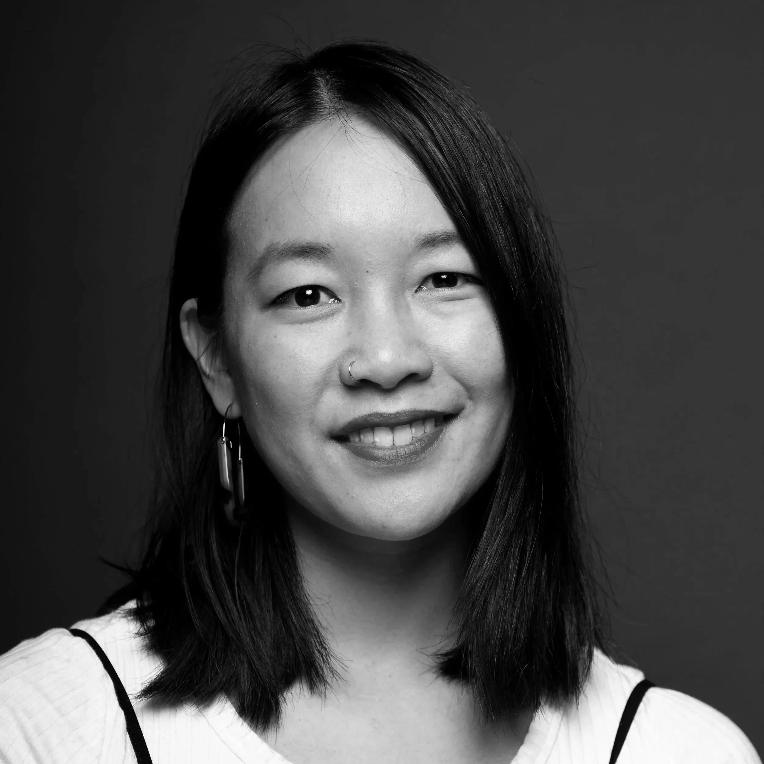 Close-up black and white portrait of an Asian woman smiling, with straight hair, wearing earrings and a white top with black straps.