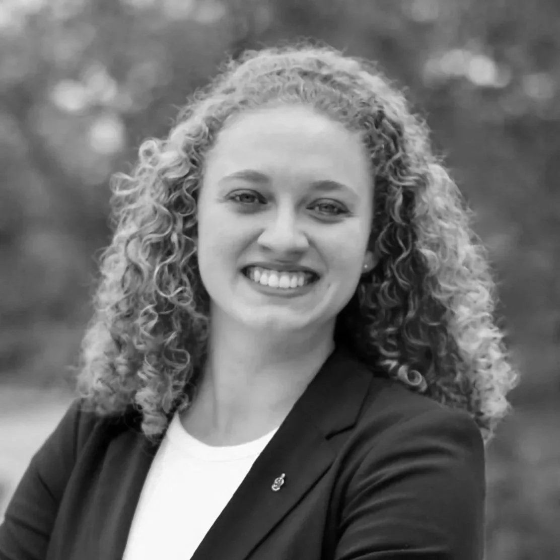 A black and white portrait of a woman with curly hair smiling and wearing a blazer.