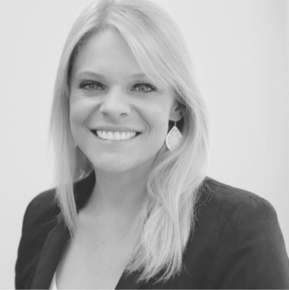 Black and white portrait of a smiling blonde woman with shoulder-length hair, wearing a blazer and a decorative earring.