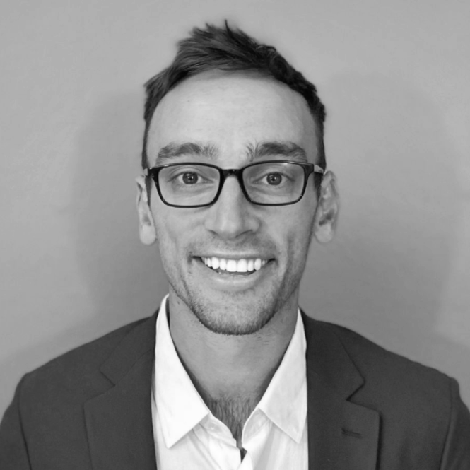 A young man with glasses, short hair, wearing a suit and tie, smiling in a black and white photo against a plain background.