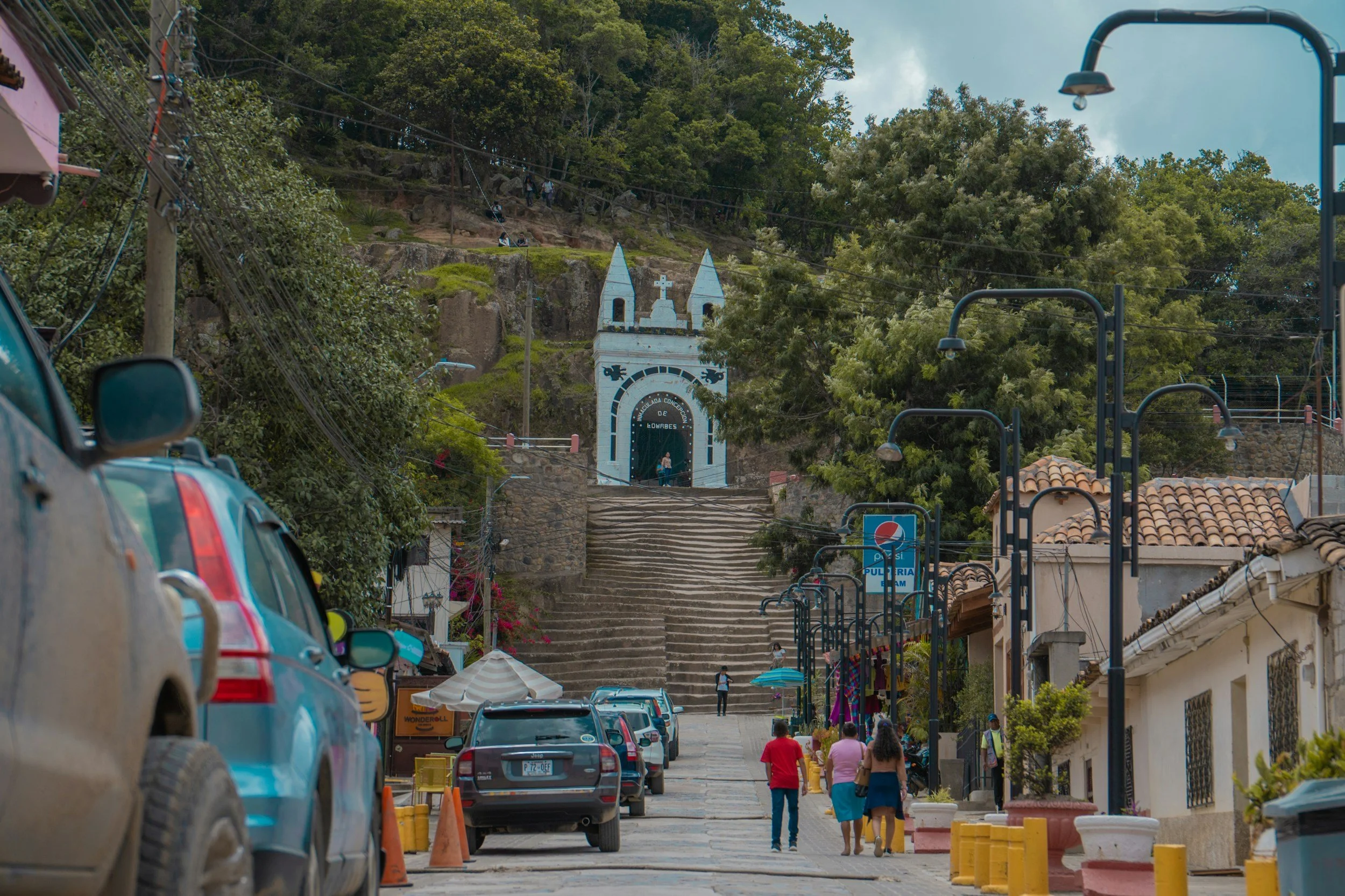 A street scene in a small town with parked cars on both sides, people walking along the sidewalk, and a church with a white tower and cross at the top at the top of a hill in the background surrounded by green trees.