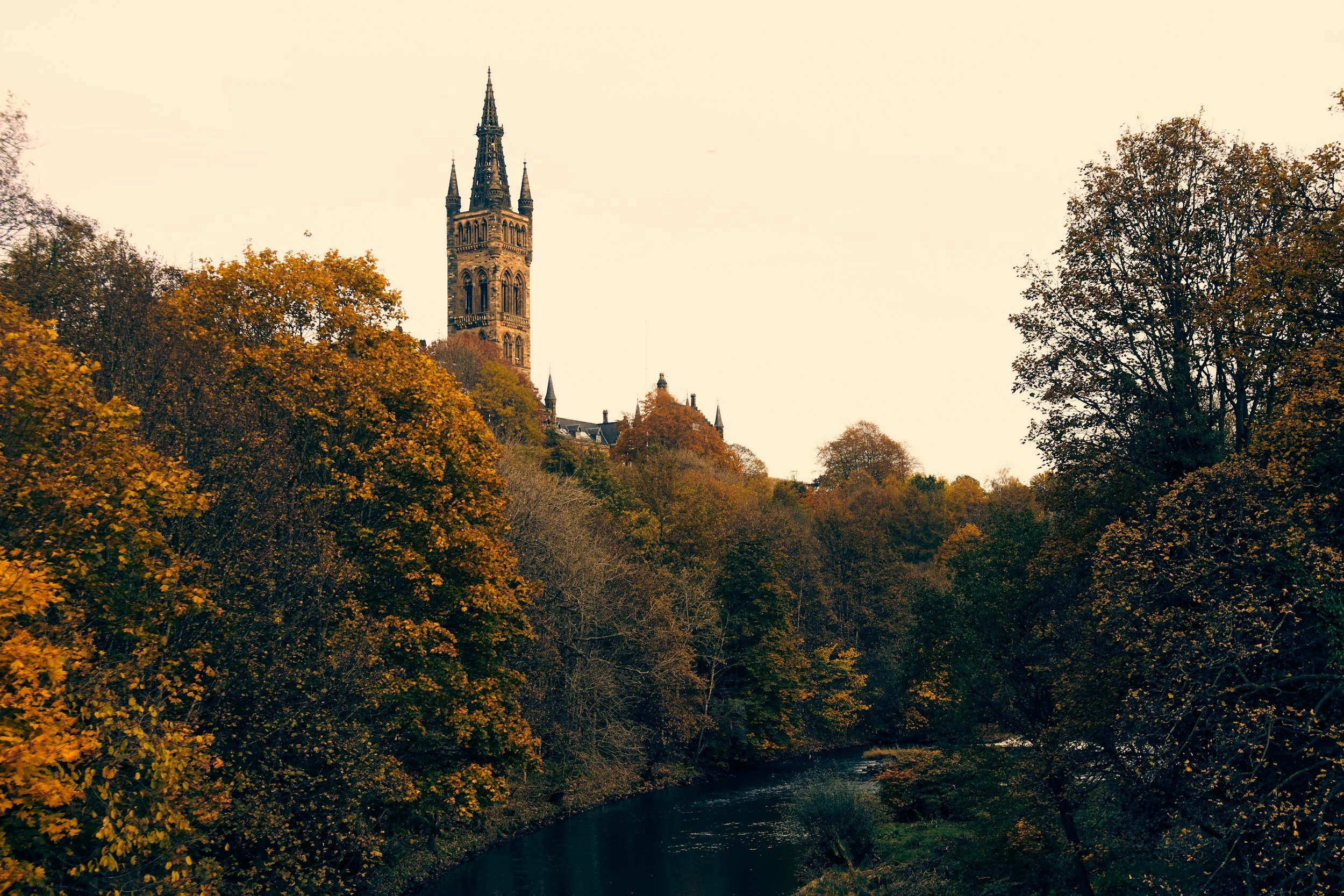 A view of a historic church tower with a tall, pointed spire, surrounded by trees with fall foliage, and a river in the foreground during daytime.