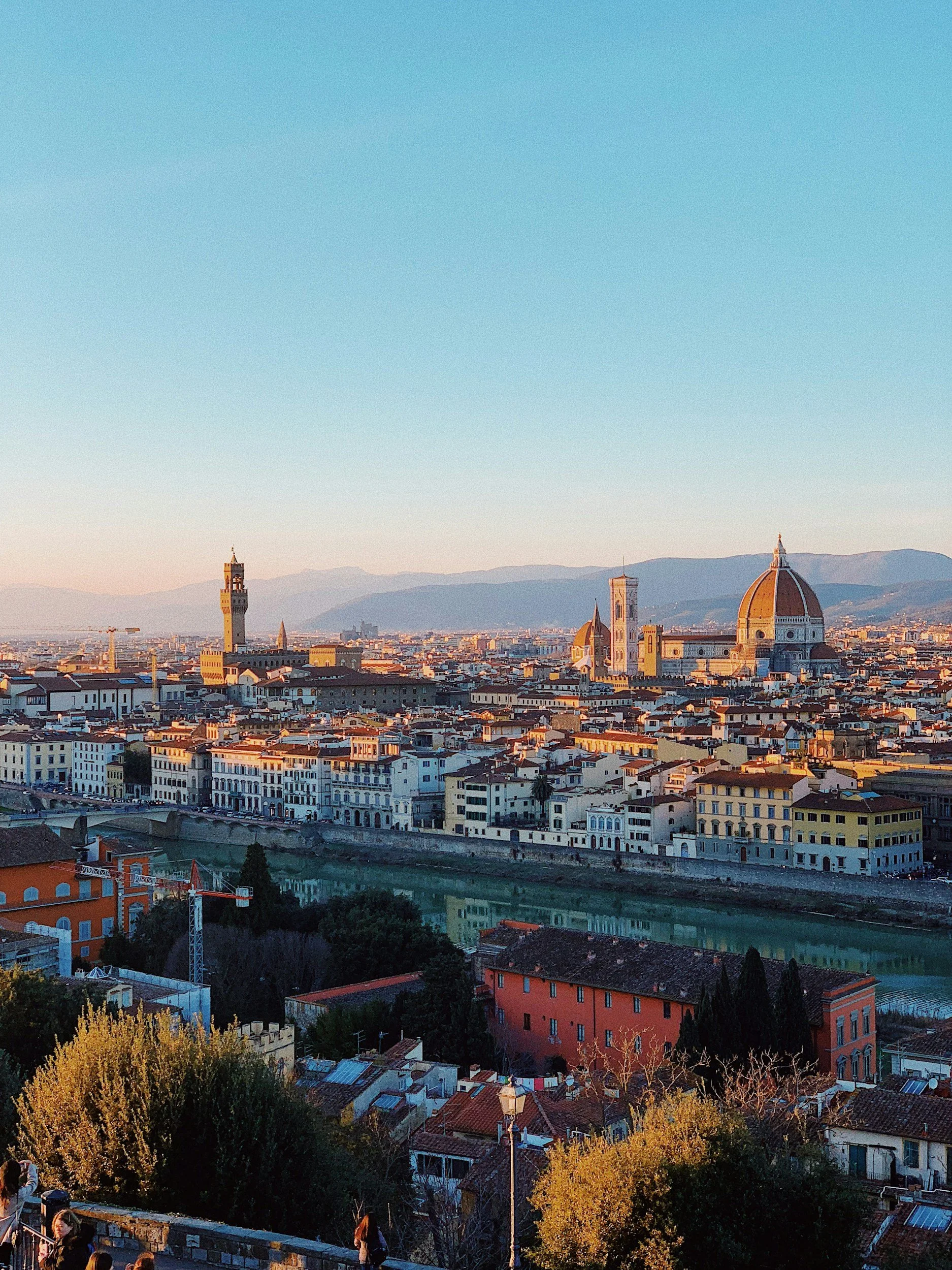 Panoramic view of Florence, Italy at sunset featuring the Cathedral of Santa Maria del Fiore with its dome, and other historic buildings and towers in the city, with mountains in the background and a clear blue sky.