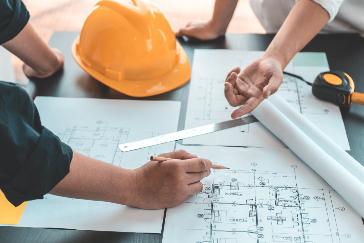 Two people review building blueprints on a table, with an orange construction helmet and measuring tools nearby.