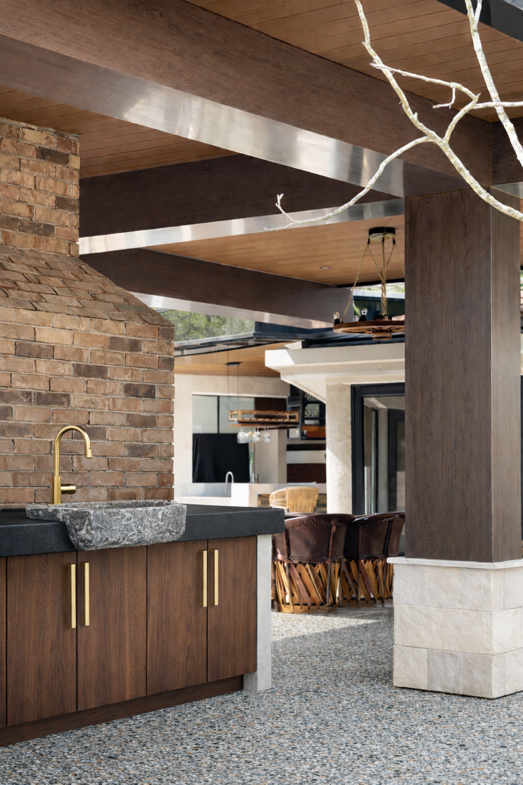 Vista del interior de una casa moderna con bloques de madera, ladrillos y mármol, incluyendo una cocina con un fregadero de piedra y un grifo dorado, y una sala con sillones de cuero. Techo con vigas de madera y construcción en estilo contemporáneo.
