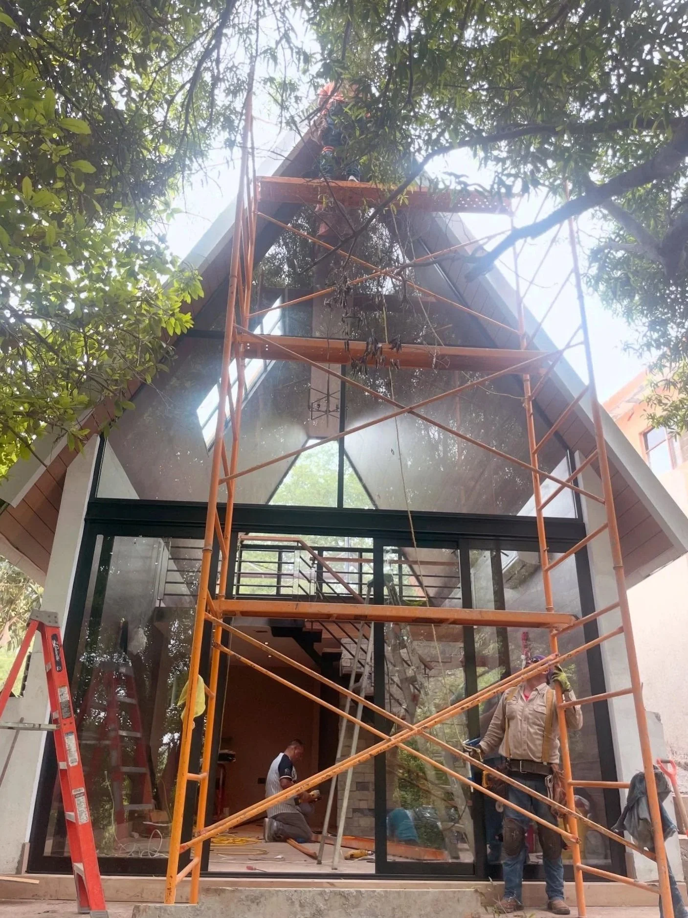 Construcción de una estructura de vidrio y metal frente a una casa moderna, con andamios naranjas y varias personas trabajando en el lugar.