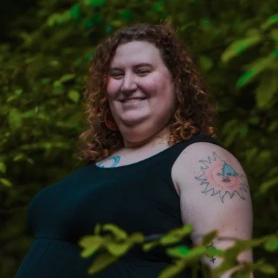 A woman with curly hair smiling outdoors, wearing a black sleeveless top, with a colorful tattoo of a sun and a face on her upper arm, surrounded by green foliage.