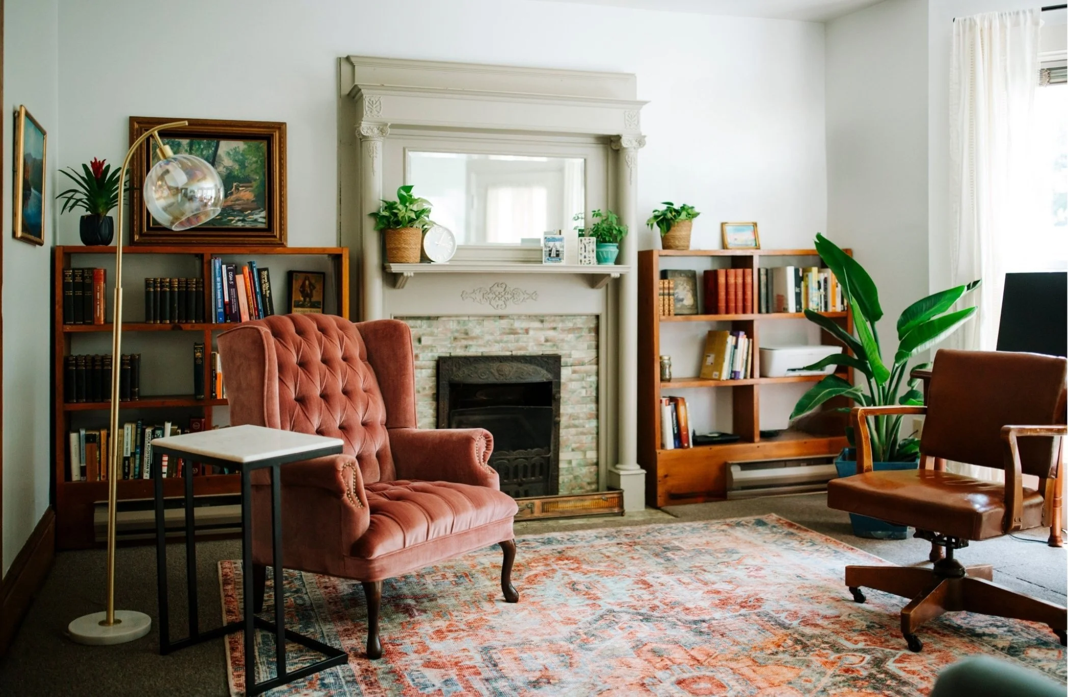 Living room with brick fireplace, pink tufted armchair, two wooden bookshelves filled with books, framed paintings, indoor plants, large mirror above fireplace, and a large window with curtains.