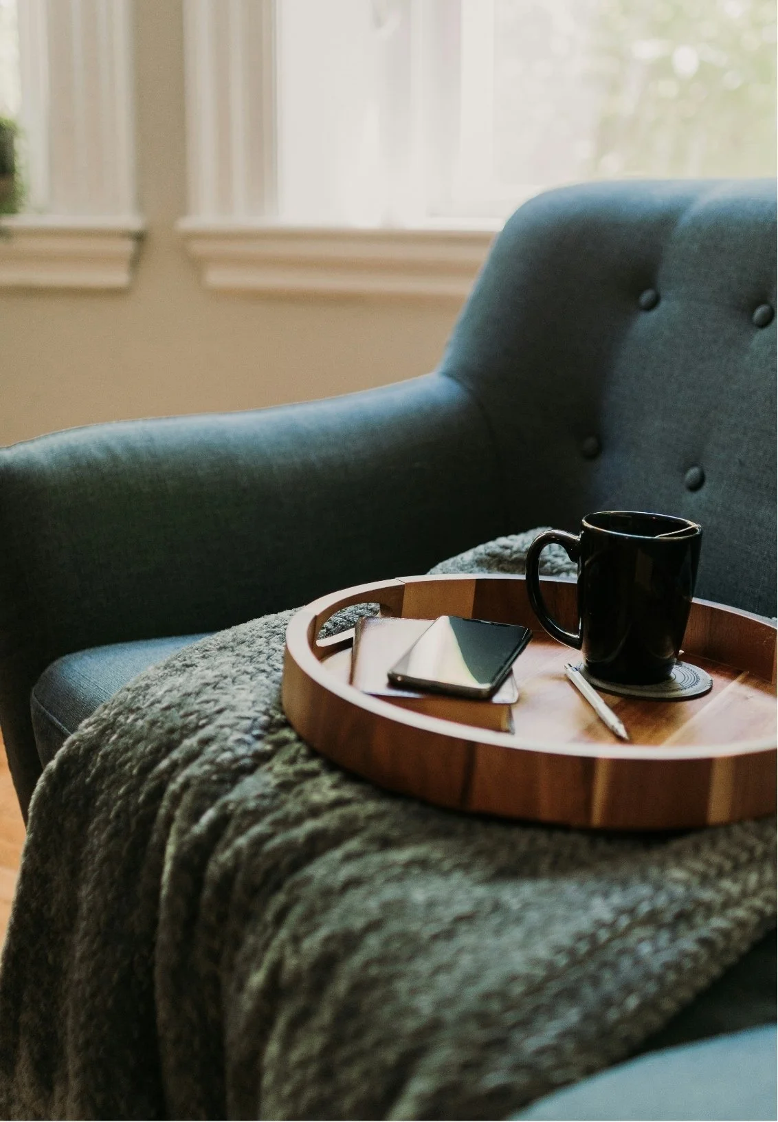 A cozy interior scene with a teal upholstered armchair, a round wooden tray, a black mug on a coaster, a closed book, and a pen on a textured blanket. Sunlight streams in through a window in the background.