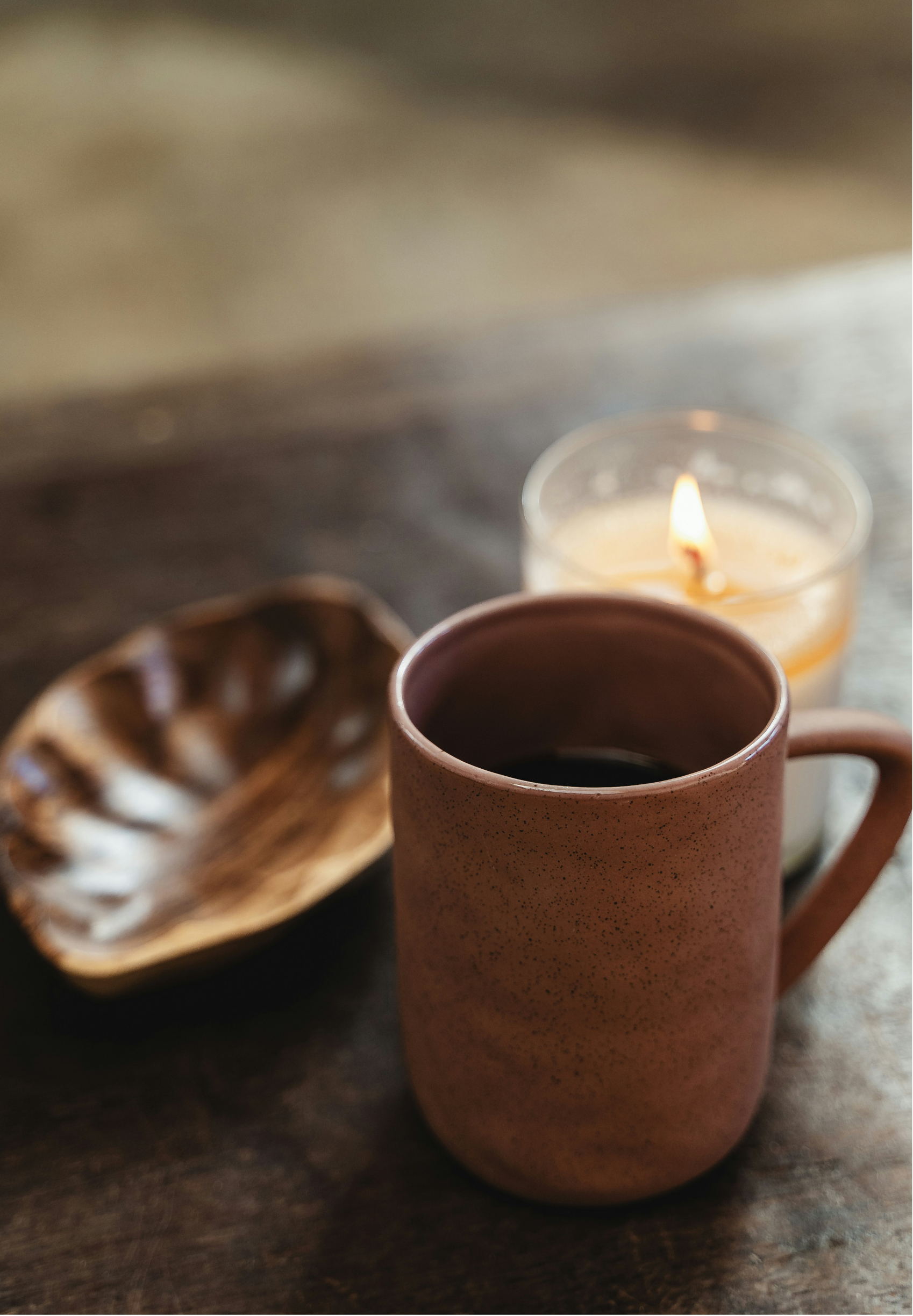 A pink speckled mug filled with black coffee, a small dish with chocolate truffles, and a lit candle in the background, all on a wooden surface.