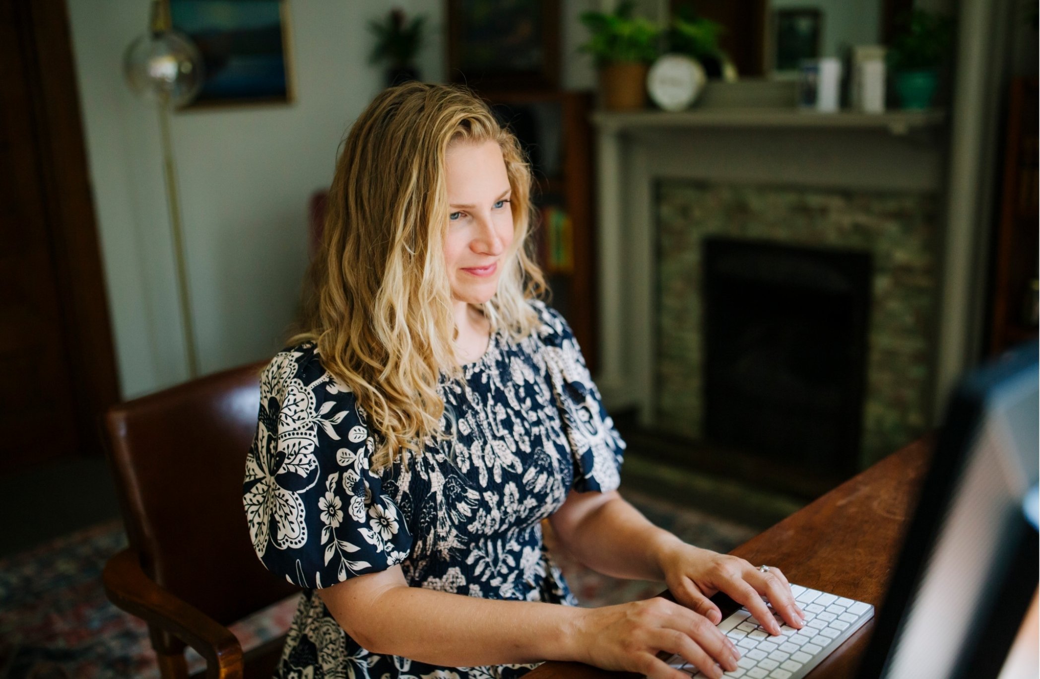Whitney Simone, psychiatrist in in Trumansburg, NY, with blonde hair typing on a keyboard in a cozy room.
