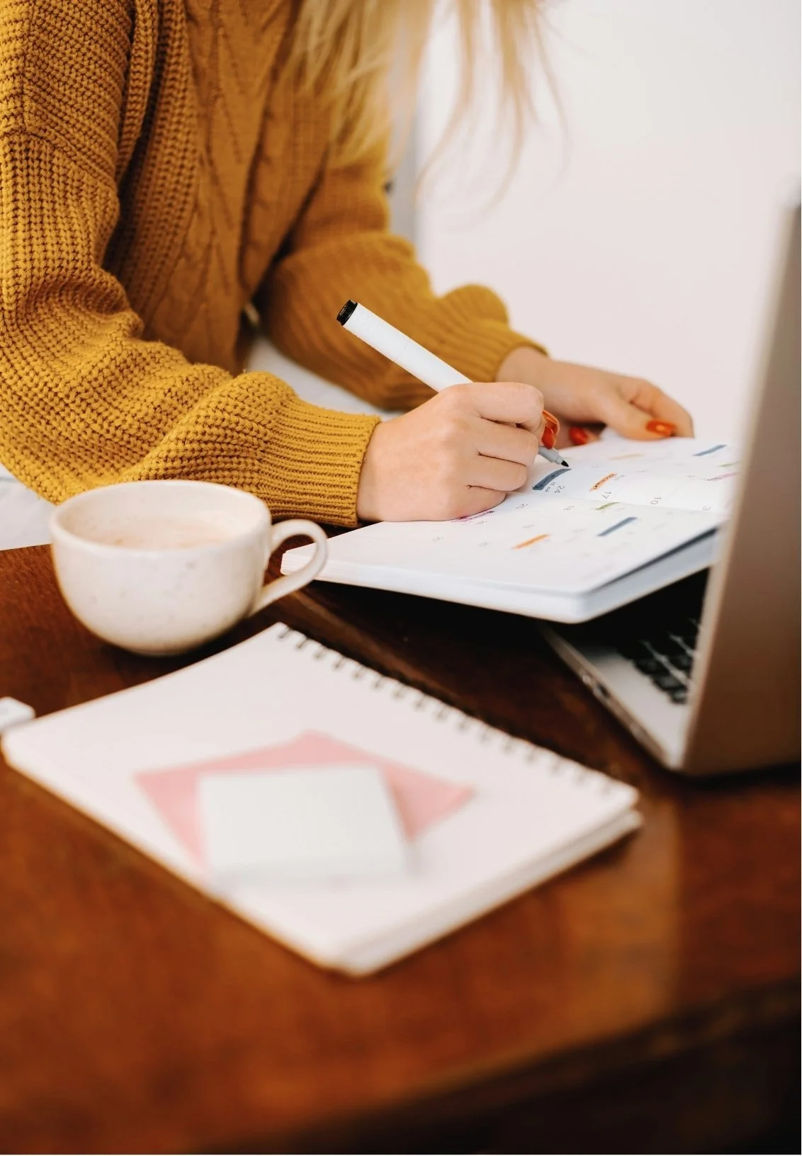 A person in a mustard yellow sweater working with a notebook and writing with a white marker, with a cup of coffee, a laptop, and a notebook on a wooden table.