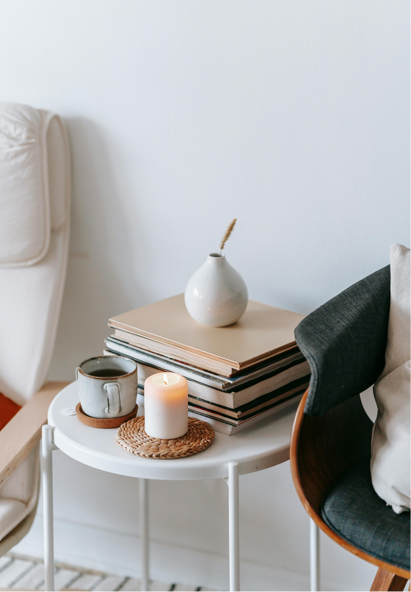 A cozy living room corner with a small round white table holding a candle, a mug of coffee, and a woven coaster, with a stack of books and a white vase with a dried flower behind it, next to a white sofa and a black armchair.