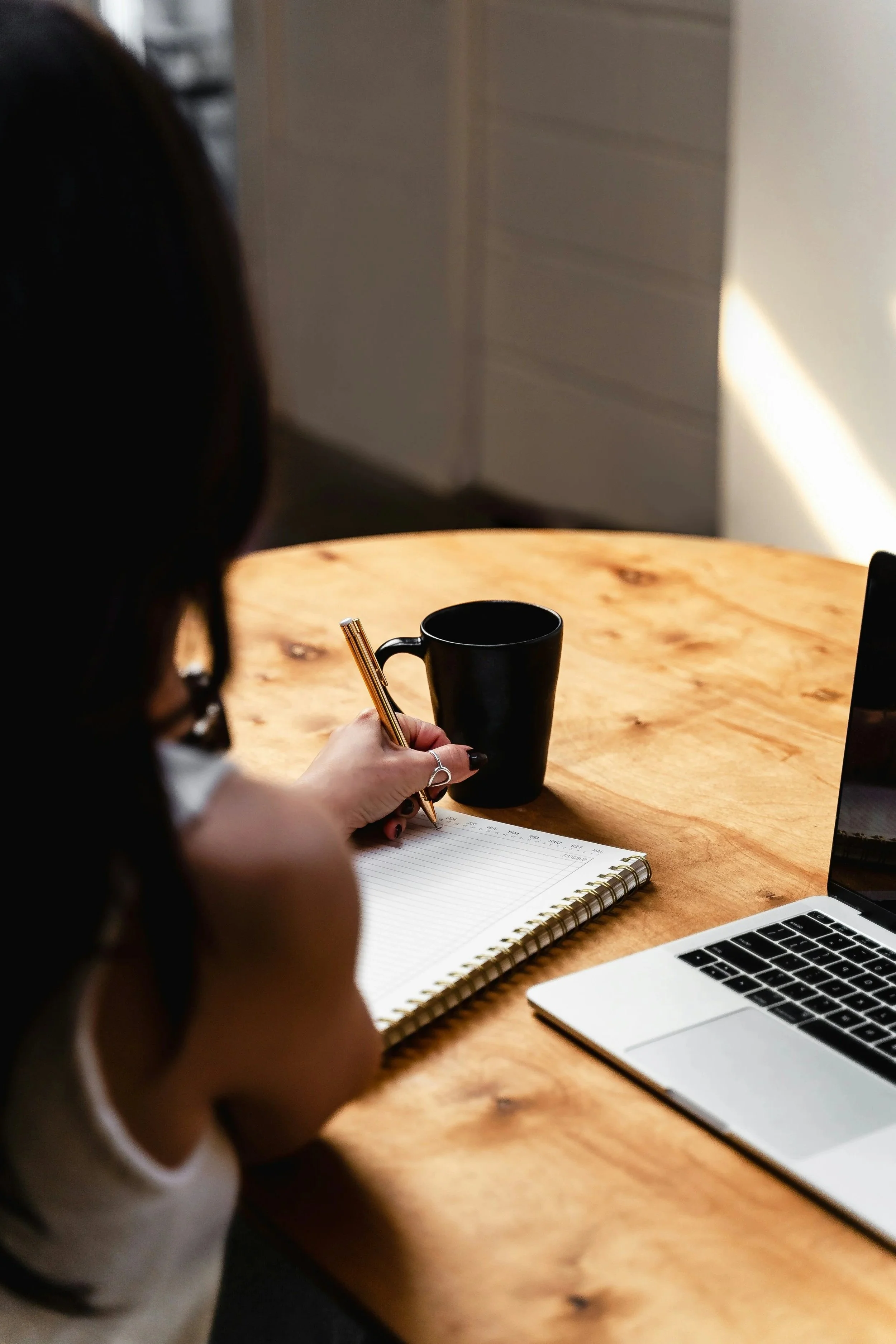 Person taking notes at a wooden table with a black mug, a laptop, and a notepad, in a well-lit room.
