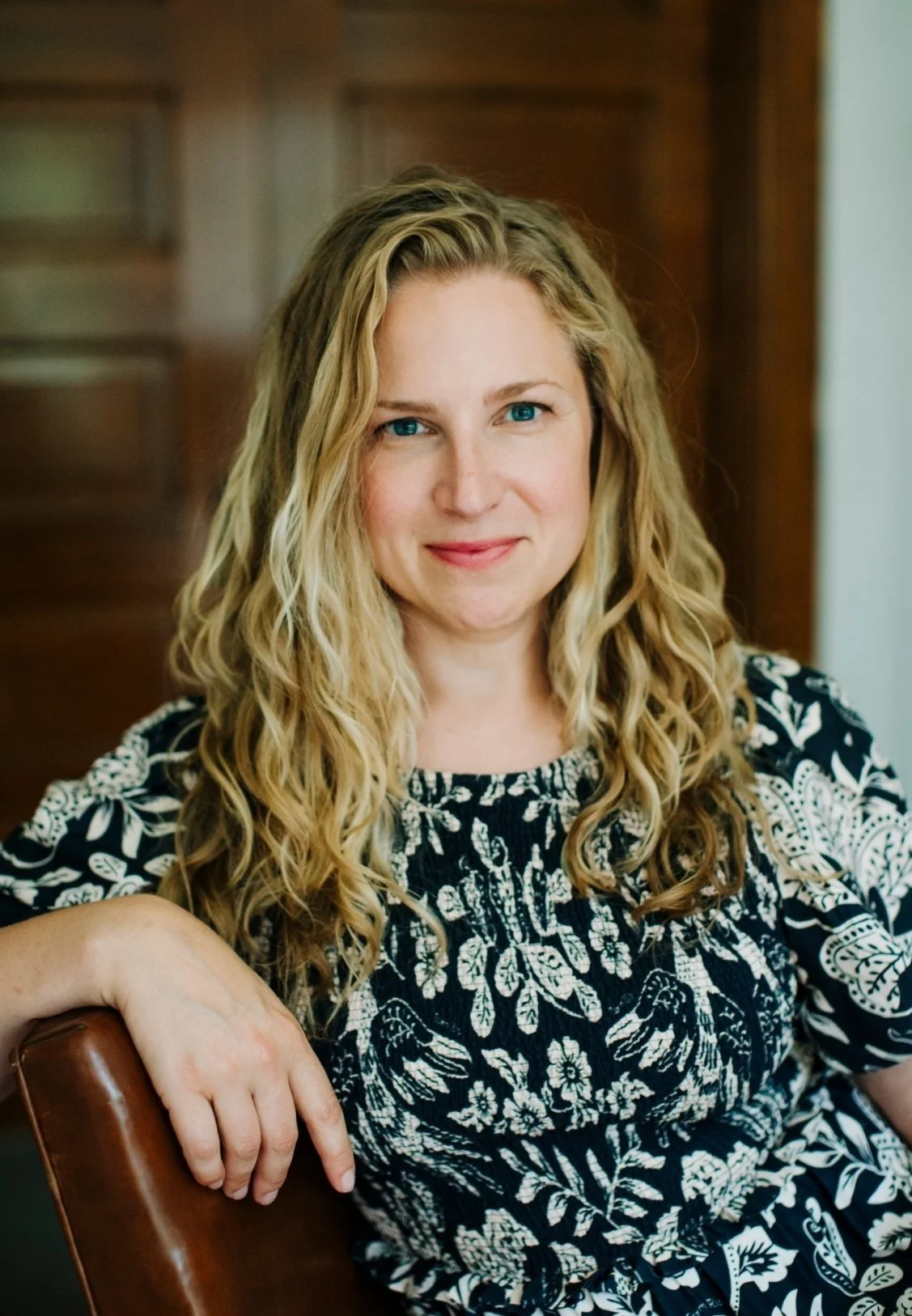 Whitney Simone, psychiatrist in in Trumansburg, NY, with blonde, curly hair and blue eyes sitting in a chair, wearing a black and white floral patterned dress, with a wooden cabinet in the background.