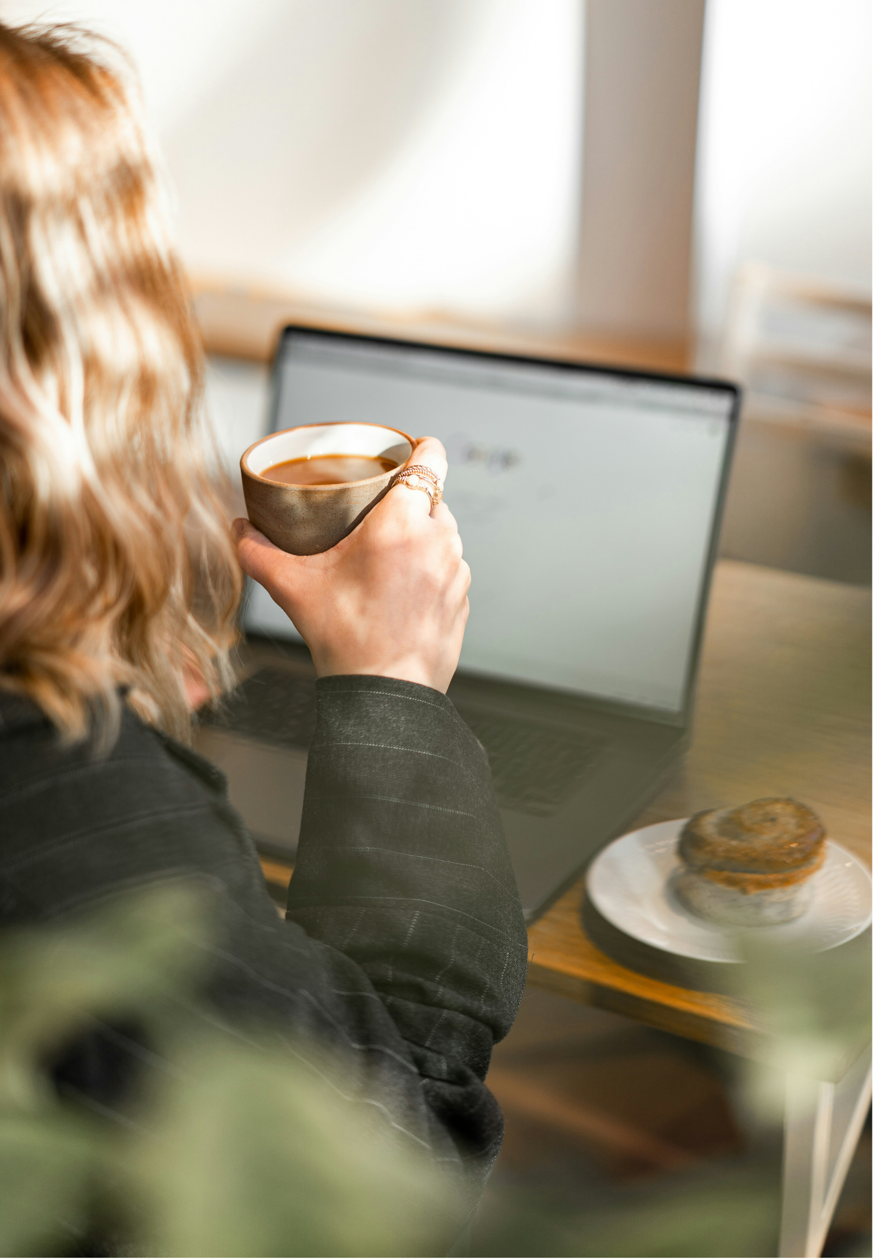 A woman with long wavy blonde hair holding a brown ceramic cup of coffee in front of a laptop, with a cinnamon roll on a plate on the desk.