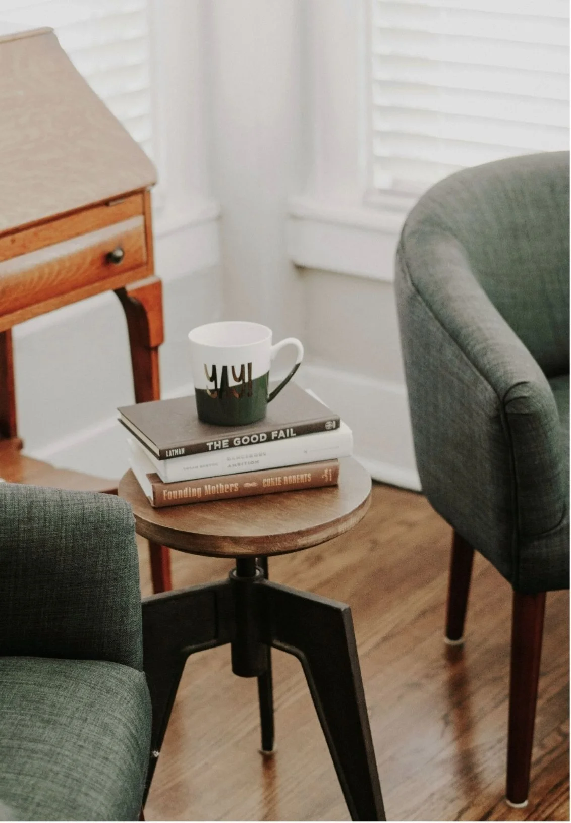 A cozy indoor scene with a round wooden side table holding a coffee mug on top of three books, surrounded by green and gray upholstered chairs, with a wooden side table, window blinds, and hardwood floor in the background.
