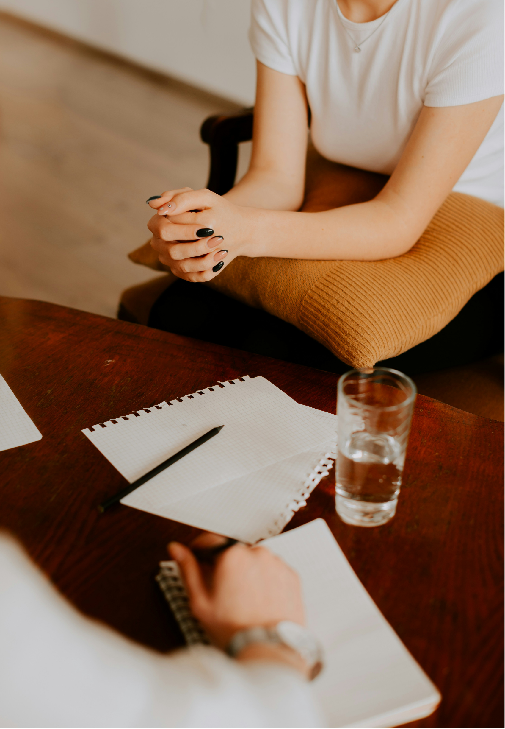 Person sitting at a table with a notepad, pen, glass of water, and a notebook, holding a pencil with hands clasped.