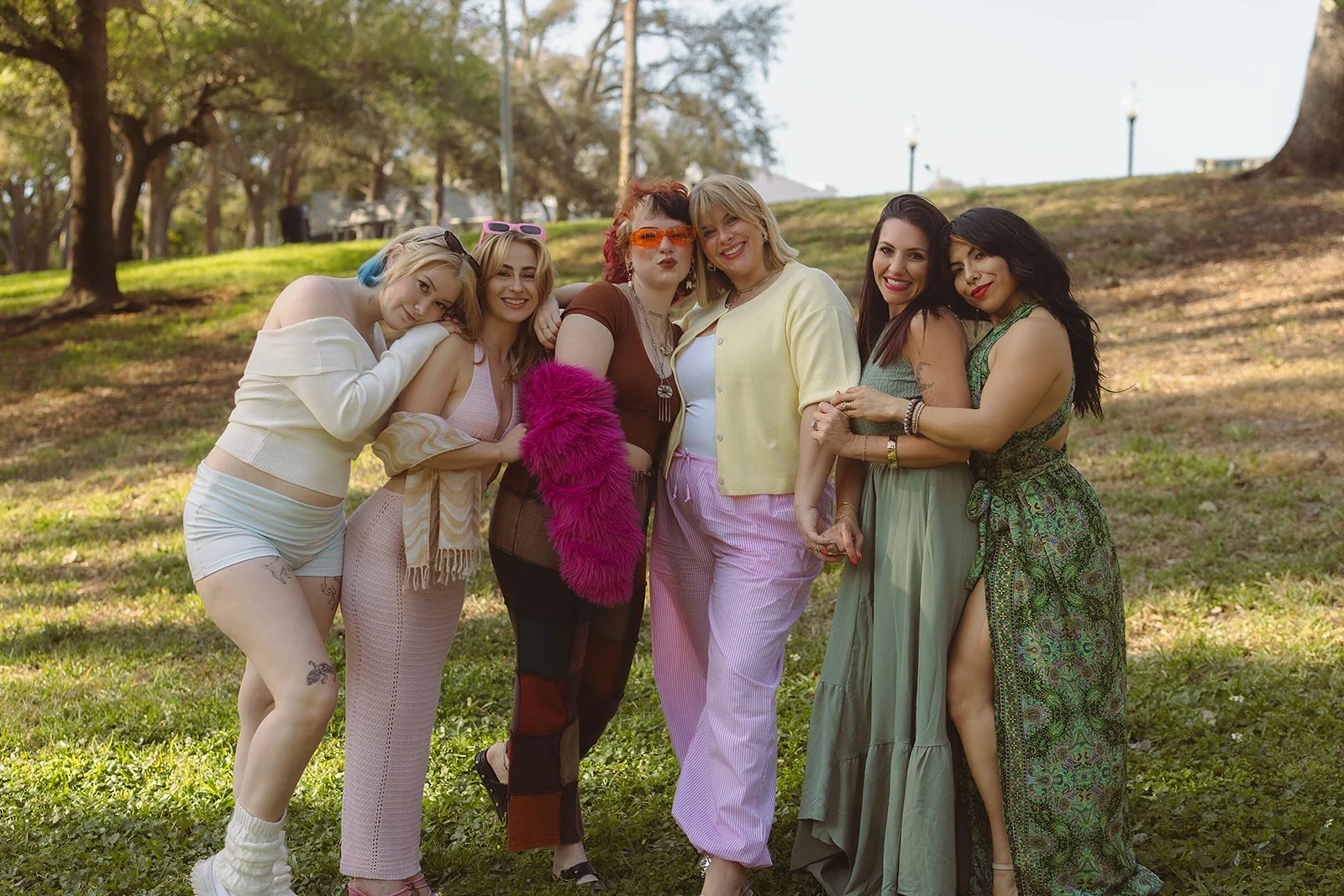 A group of six women standing close together outdoors on a grassy area with trees and lampposts in the background, smiling and hugging each other.