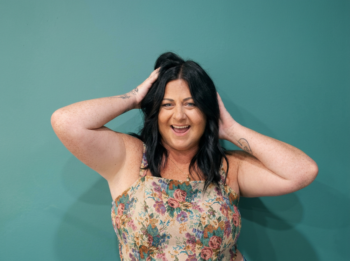 A woman with black hair, tattoos, and freckles smiling and touching her hair against a teal wall.