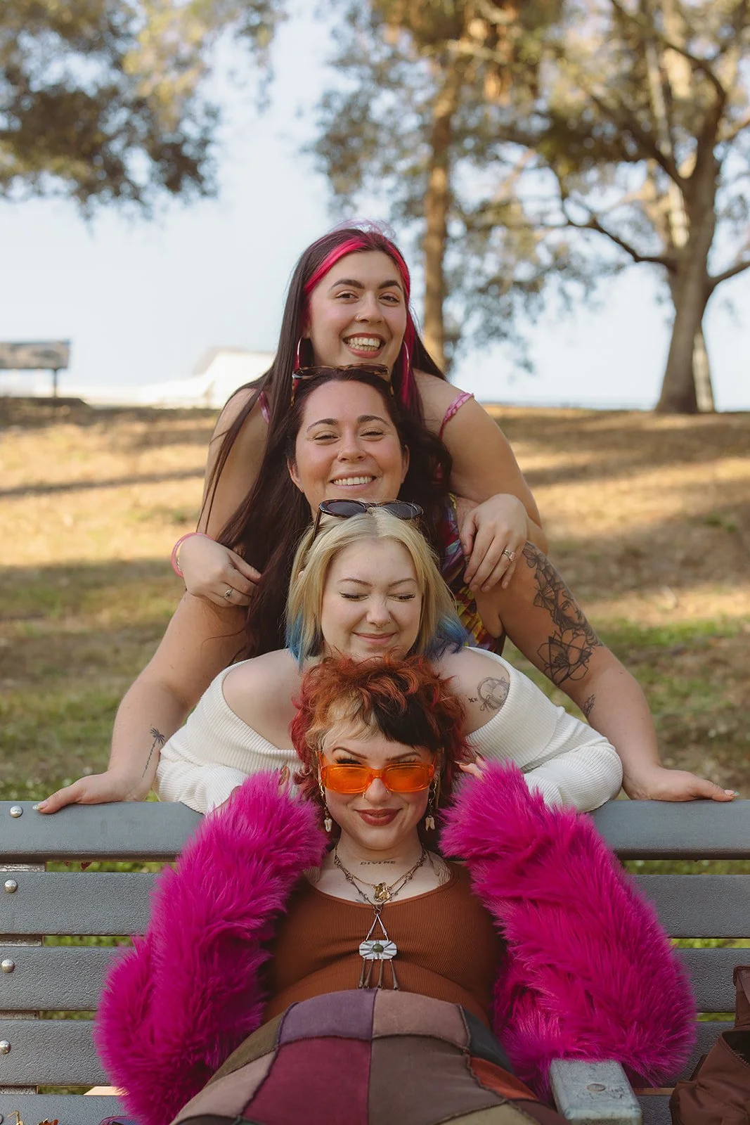Five women with colorful hair and accessories, smiling and smiling on a park bench and in a line, with trees and a clear sky in the background.