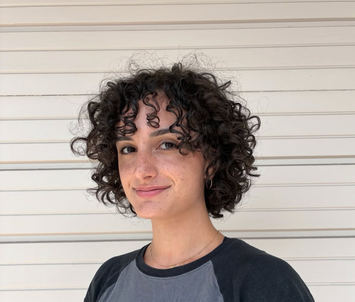 A young woman with short, curly dark hair, light skin with freckles, smiling, wearing a black and gray top, standing against a white horizontal panel wall.