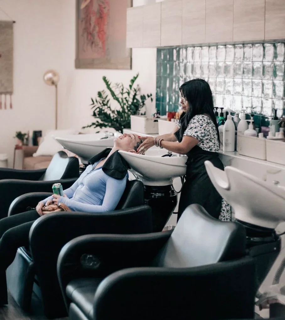 A woman receiving a hair wash at a salon, seated with her head tilted back over a washing sink, while a stylist shampoos her hair.