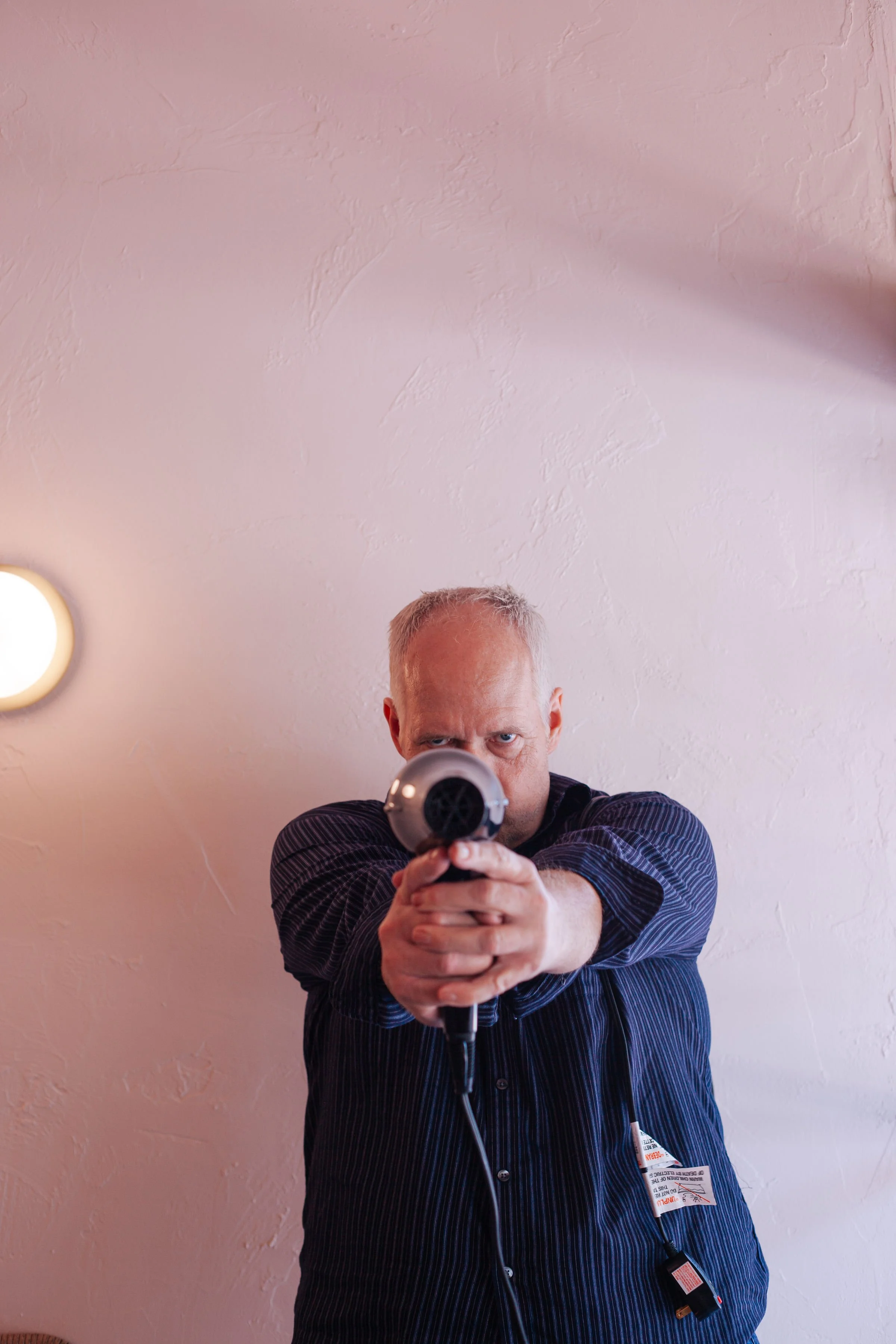 A man with short gray hair, wearing a black striped shirt, is pointing a gun at the camera. He is standing against a textured pink wall with a round light fixture on the left side of the wall.