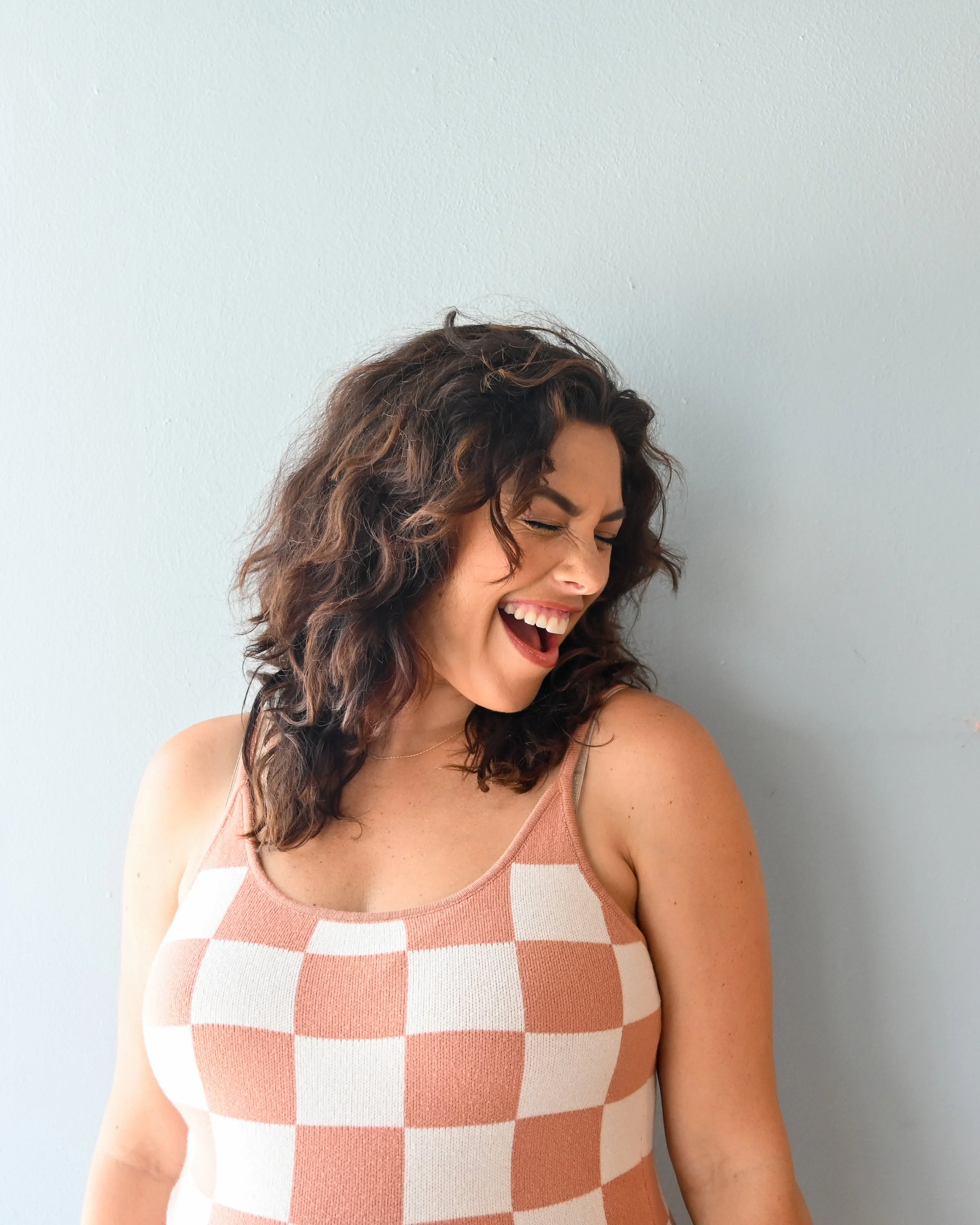 A woman with curly brown hair is smiling and laughing, wearing a sleeveless dress with a checkered pattern in shades of coral and white, standing against a plain, light-colored wall.