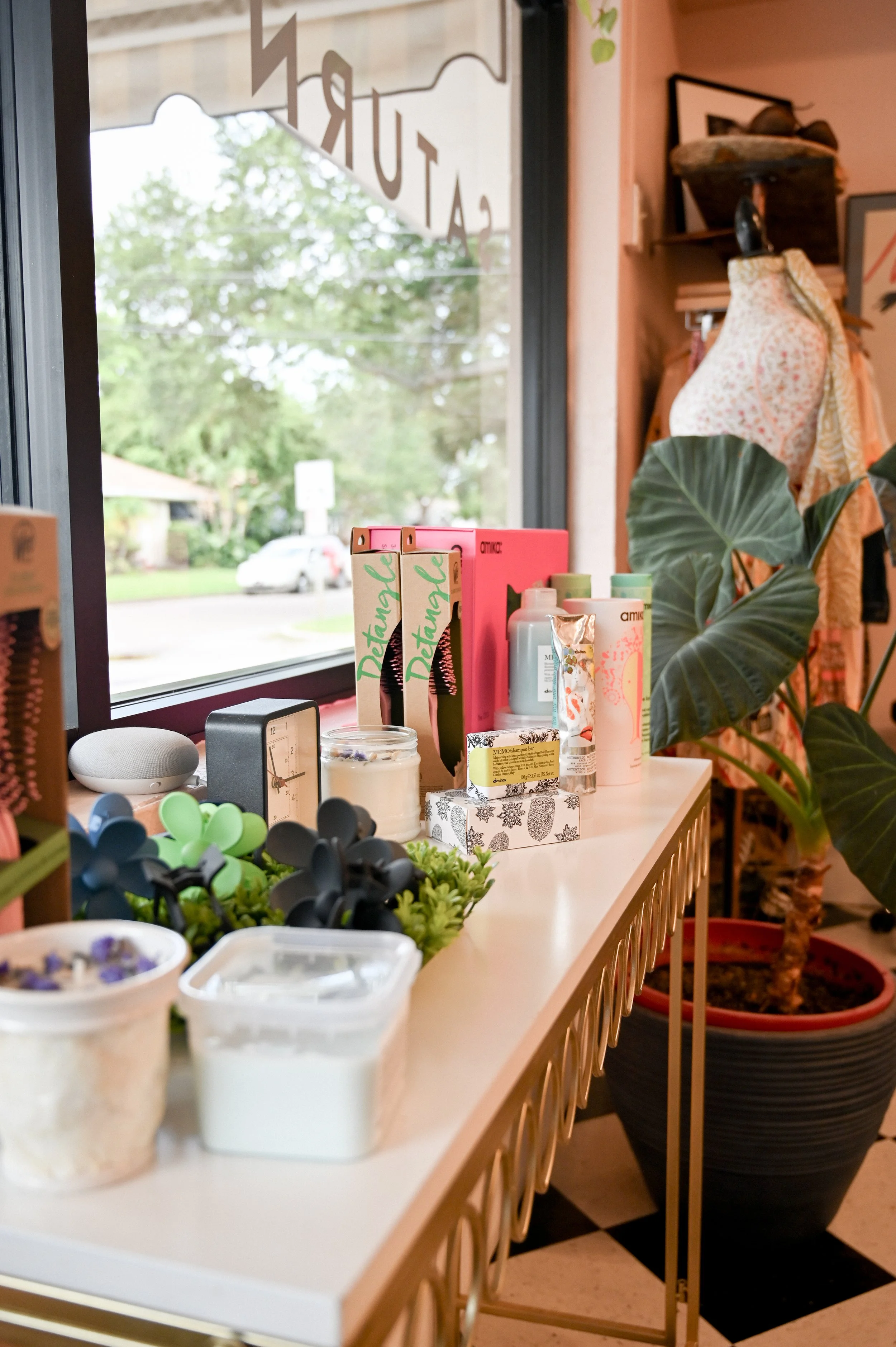 Display inside a store with various skincare products, candles, and decorative items on a white table near a window with green trees outside.
