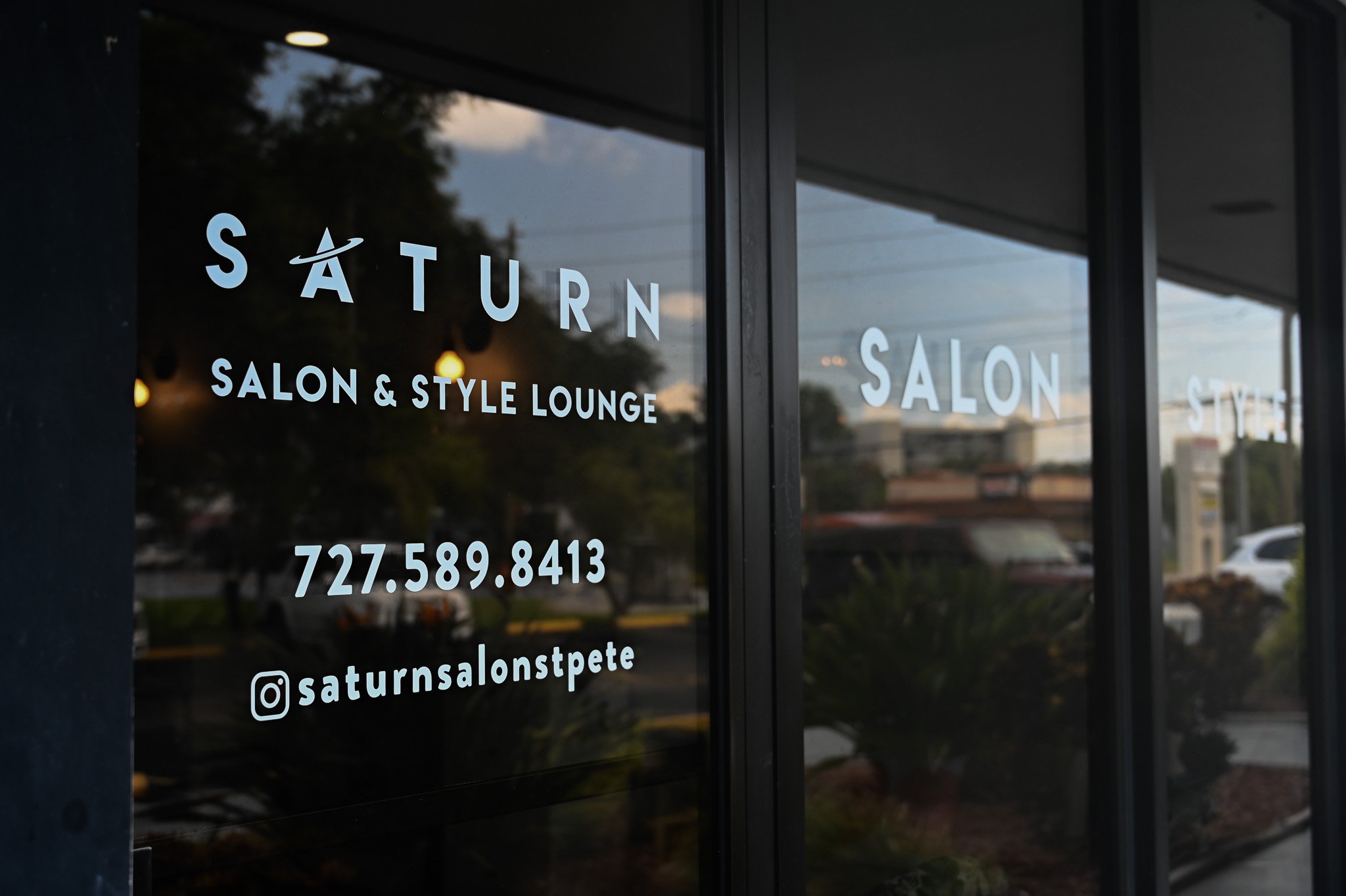 The glass window of a salon with the name 'Saturn Salon & Style Lounge' and a phone number. The reflection of trees and buildings can be seen in the glass.