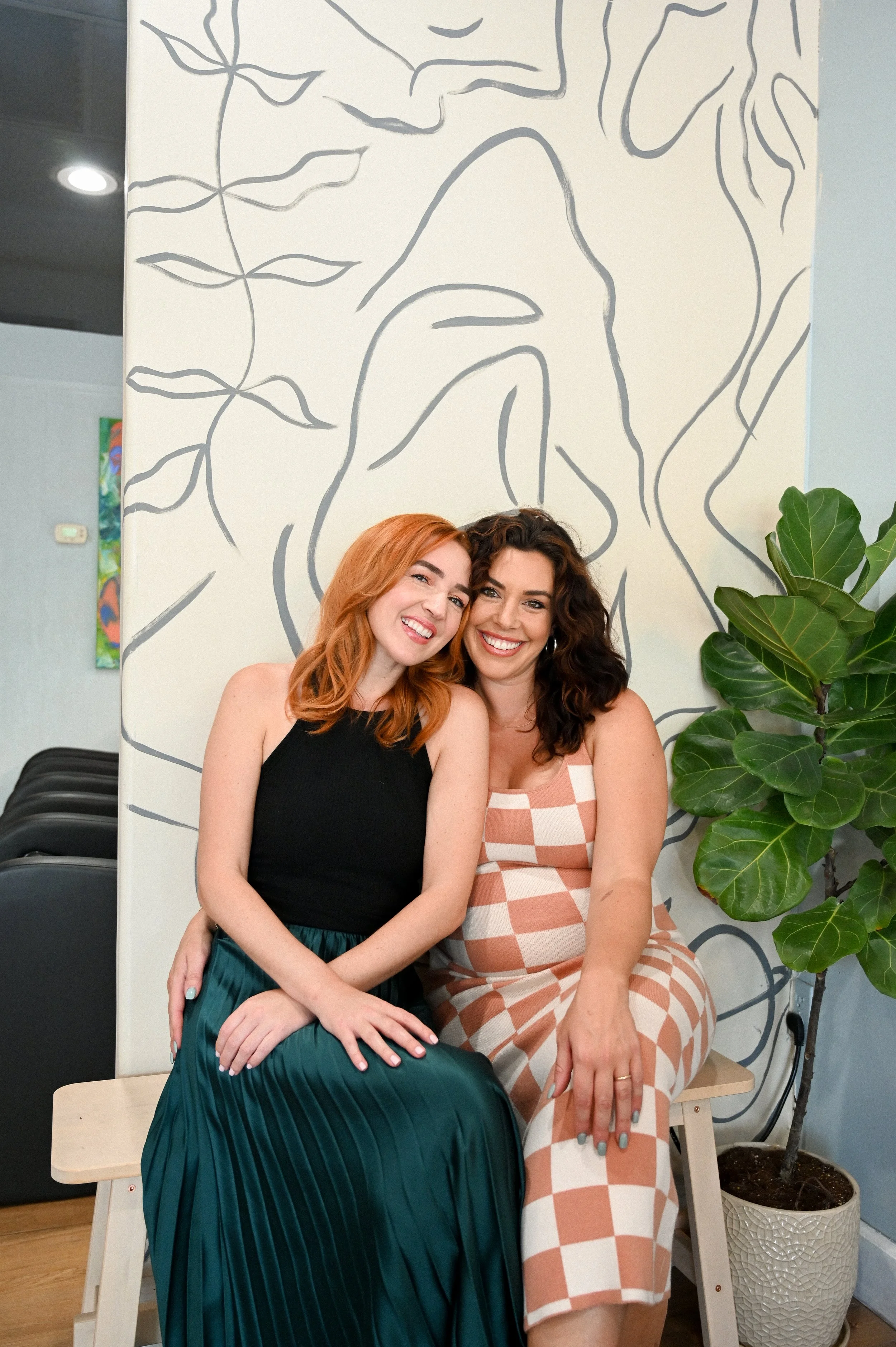 Two women sitting closely together, smiling, in front of a white wall with abstract black line art and a potted plant.