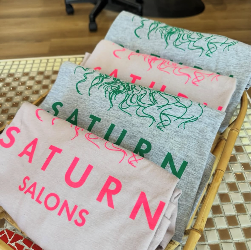 Four folded T-shirts with different designs and colors, laid out in a basket on a table. The T-shirts have patterns and text related to Saturday, Sunday, and salons.