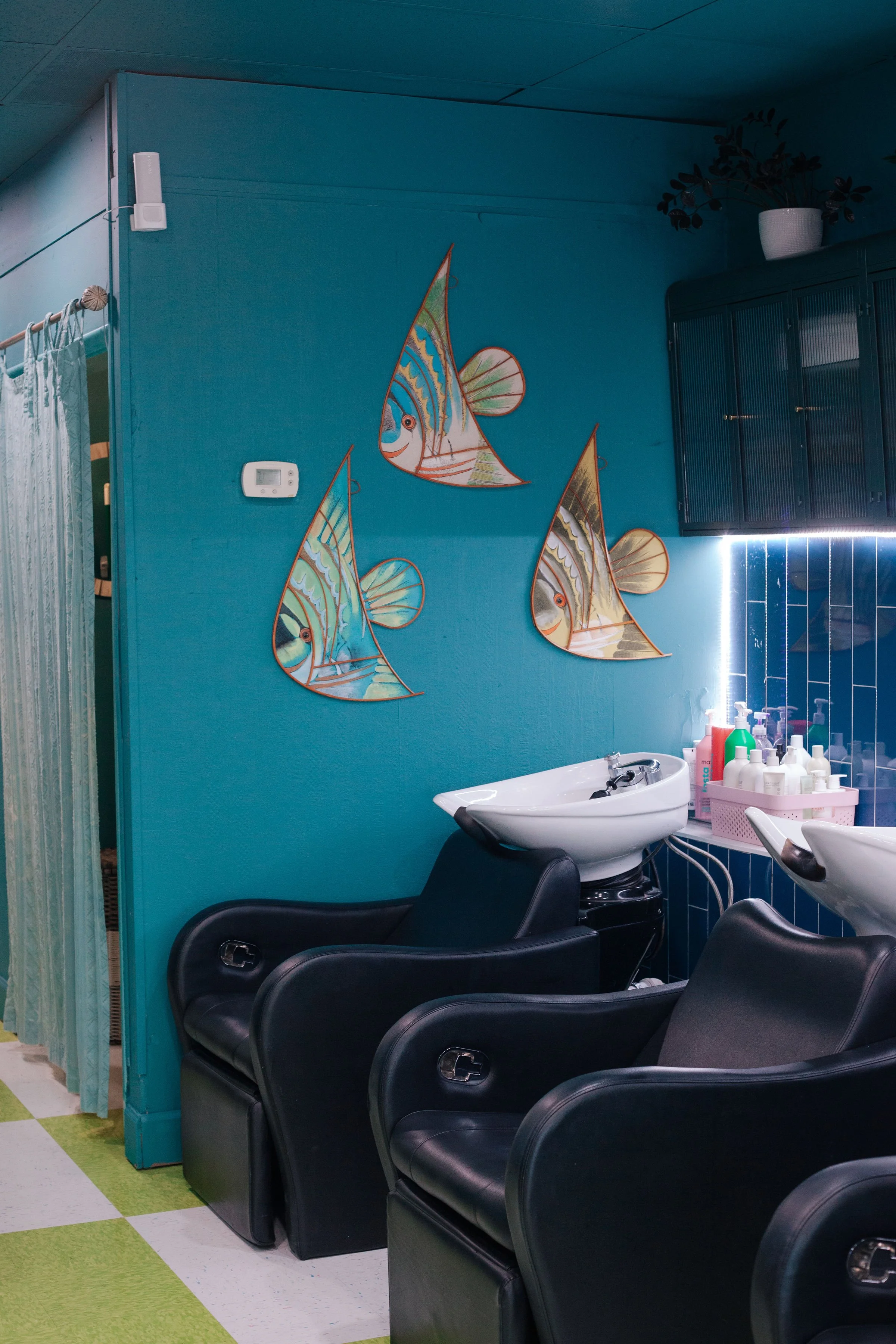 A salon with two black barber chairs in front of white sinks. On the wall, there are three colorful fish-shaped decorations. To the right, there are various bottles and hairdressing supplies on a white cart, and a blue cabinet with a potted plant on 