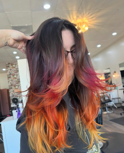 Woman with long, multi-colored hair in shades of black, purple, pink, orange, and yellow inside a hair salon, wearing glasses and touching her hair.
