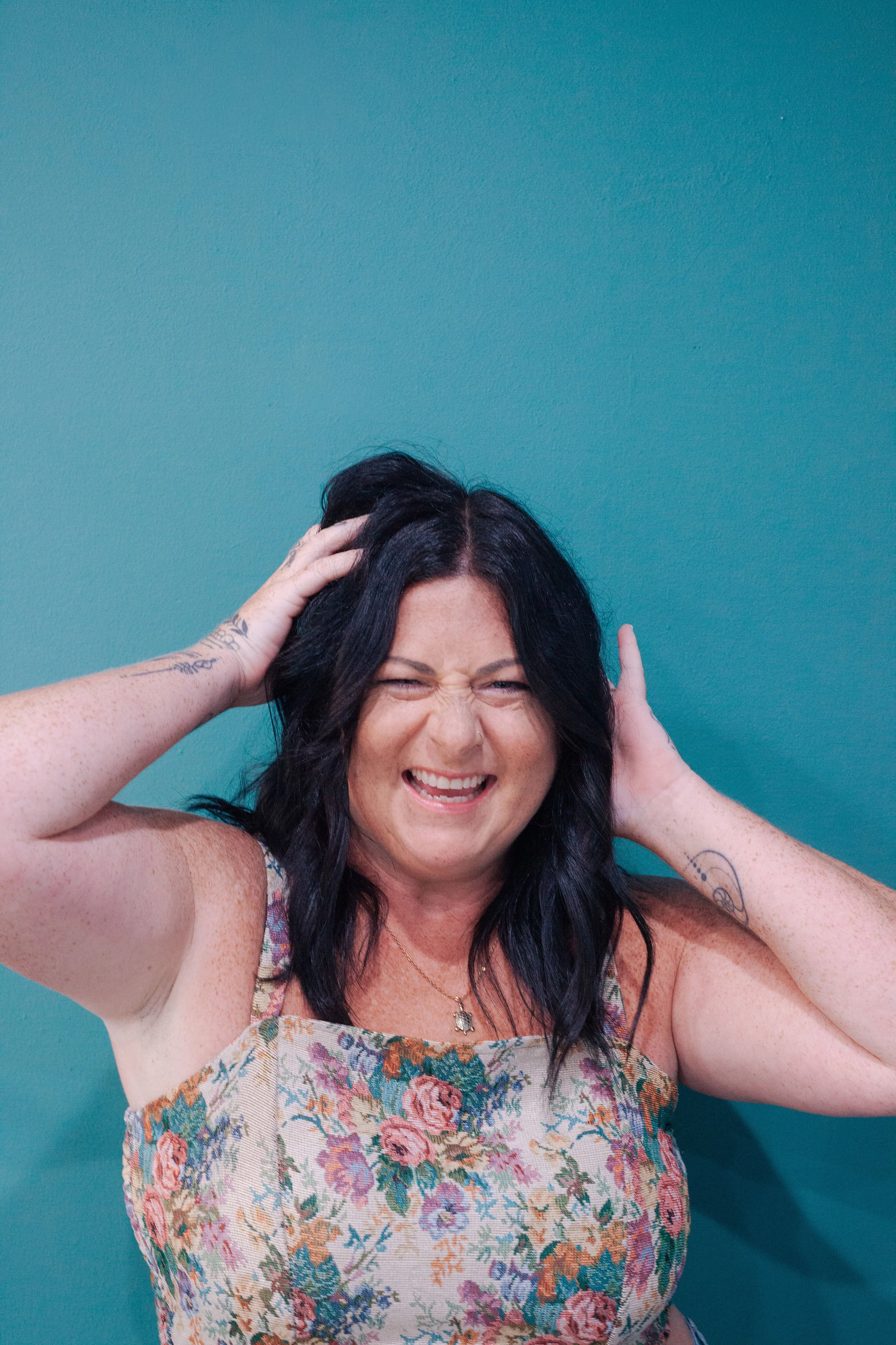 A woman with long dark hair smiling widely, holding her head with both hands, wearing a floral dress, in front of a teal wall.