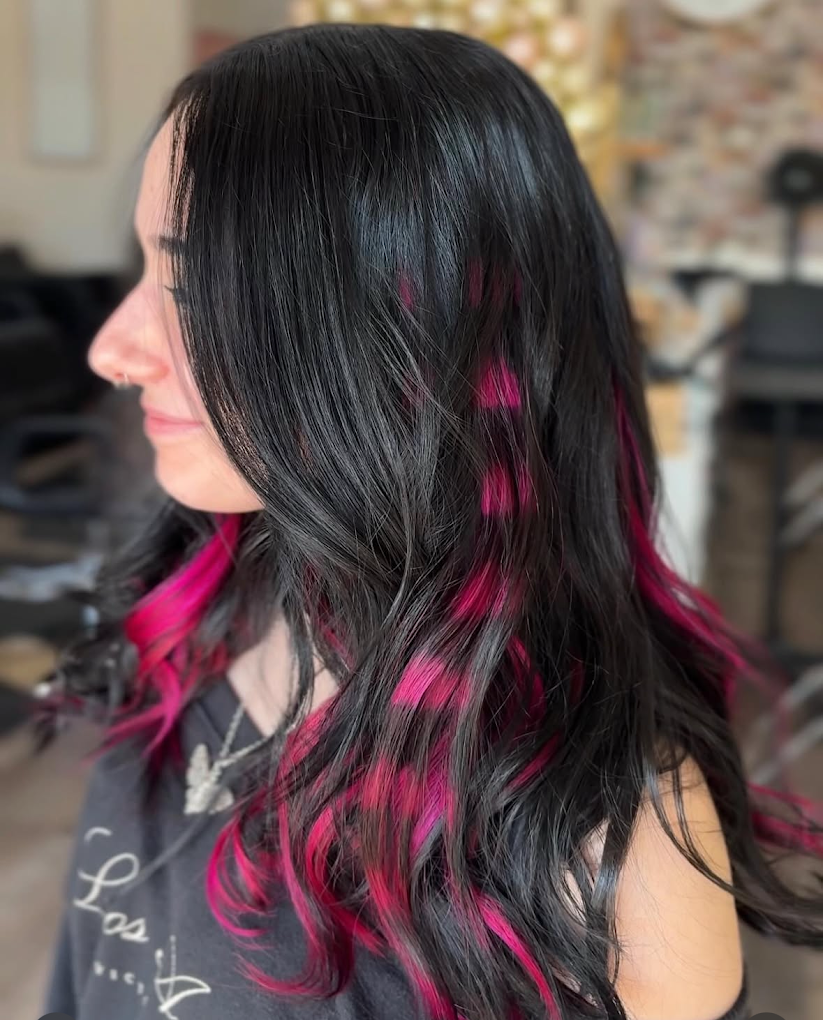 Side view of a woman with long black hair with pink highlights, sitting in a hair salon.