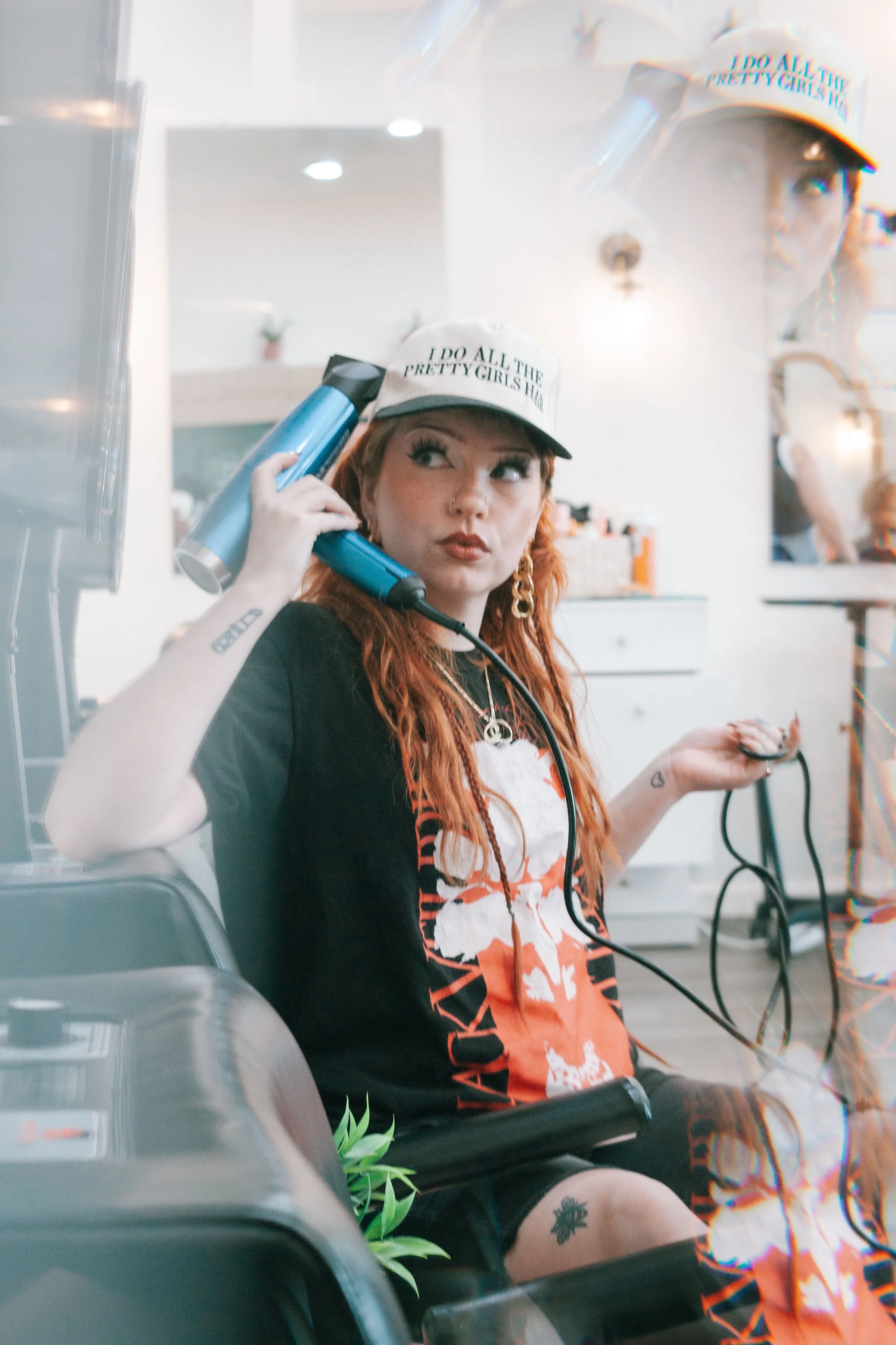 Young woman with red hair and tattoos, holding a hairdryer, sitting in a salon, wearing a cap that reads 'I DO ALL THE PRETTY GIRLS HAIR', and looking into a mirror.