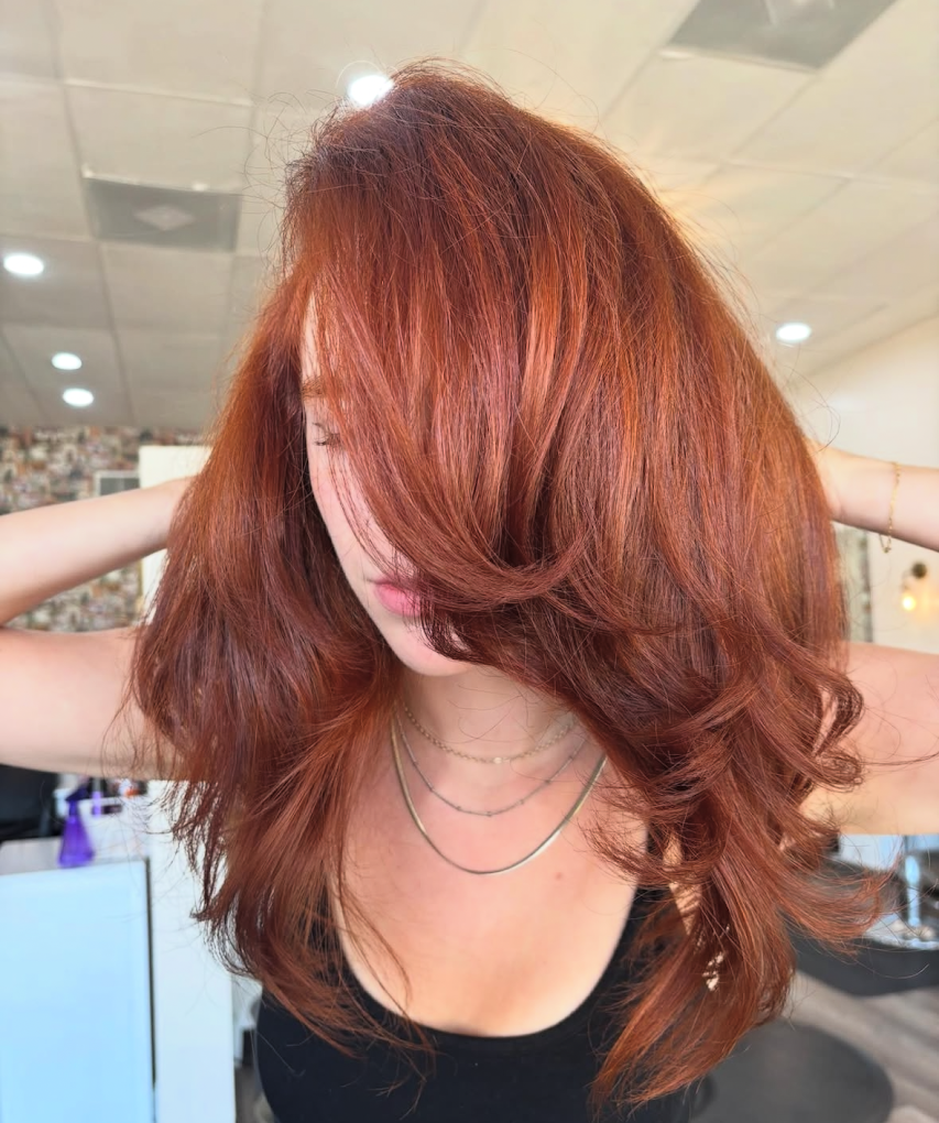 A woman with long, voluminous red hair covering part of her face, wearing layered necklaces, with her arms raised behind her head in a salon or studio setting.