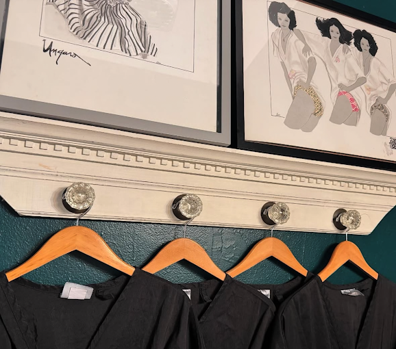 Four empty wooden hangers hanging on a white-painted nail rack, with black garments draped over them, below framed fashion illustrations on a teal wall.