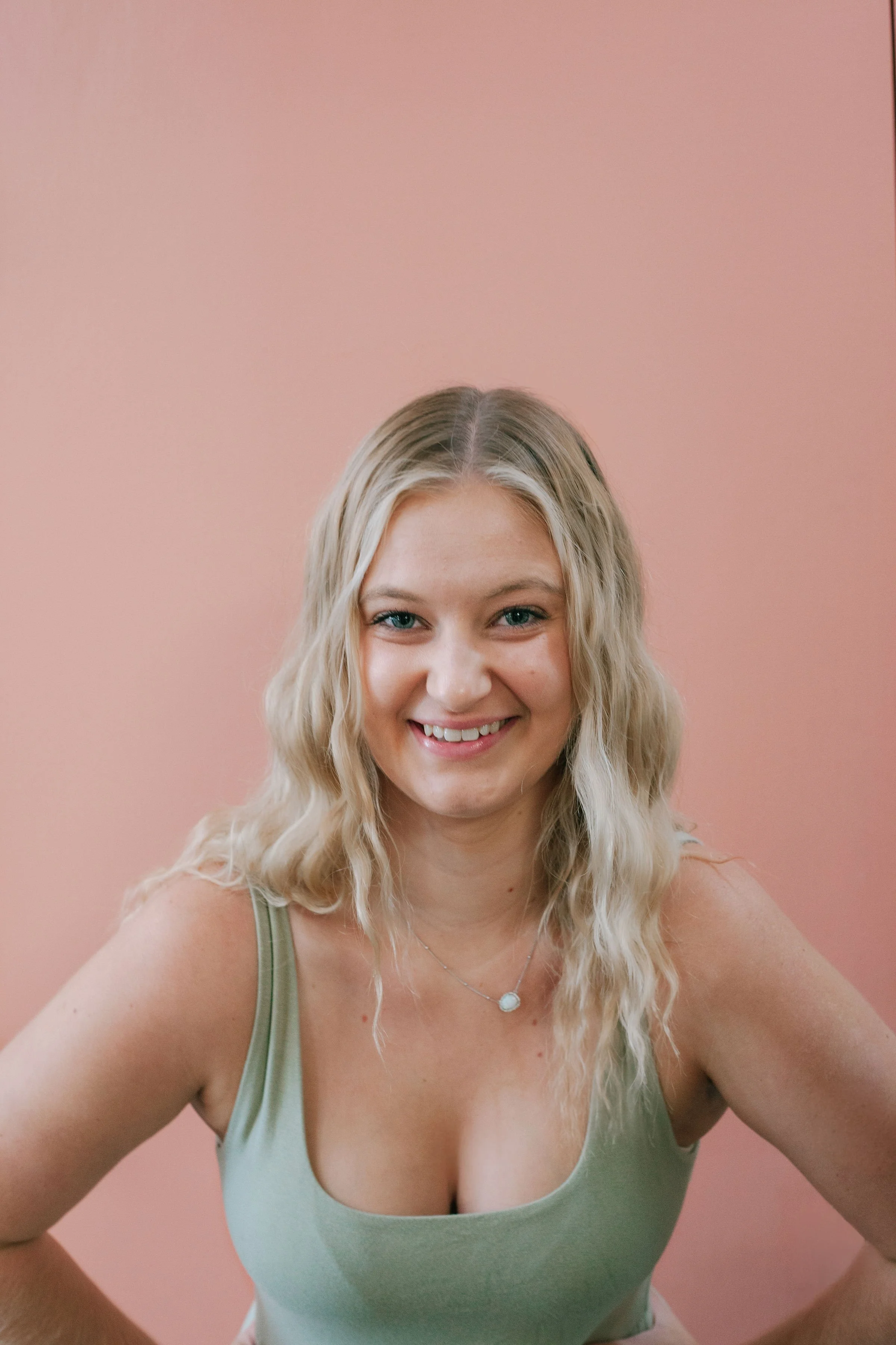 A young woman with blonde, wavy hair taking a selfie smiling at the camera against a pink background.