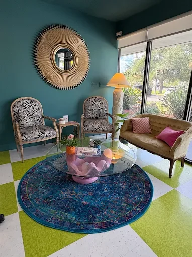 A colorful waiting area with two patterned armchairs, a pink sofa, a glass coffee table, and a blue round rug. A large sunburst mirror hangs on a teal wall, with a window showing outdoor trees and a street.