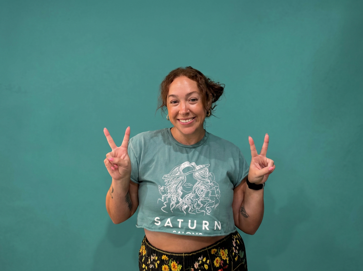 A smiling woman with curly hair making peace signs with both hands, standing against a teal background, wearing a cropped teal T-shirt with a graphic of a woman with a hat and the word 'SATURN' and a black floral skirt.