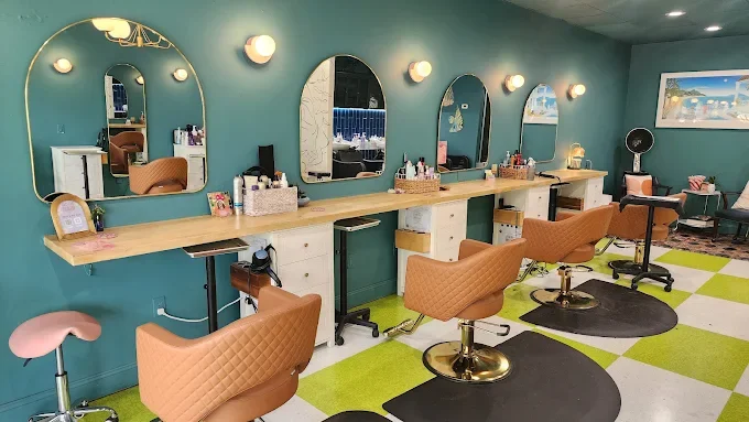 Beauty salon with four styling stations, each with a mirror, light, and chair, on a wall painted teal. The floor has green and white checkerboard flooring, and there are additional seating and decoration towards the back of the room.