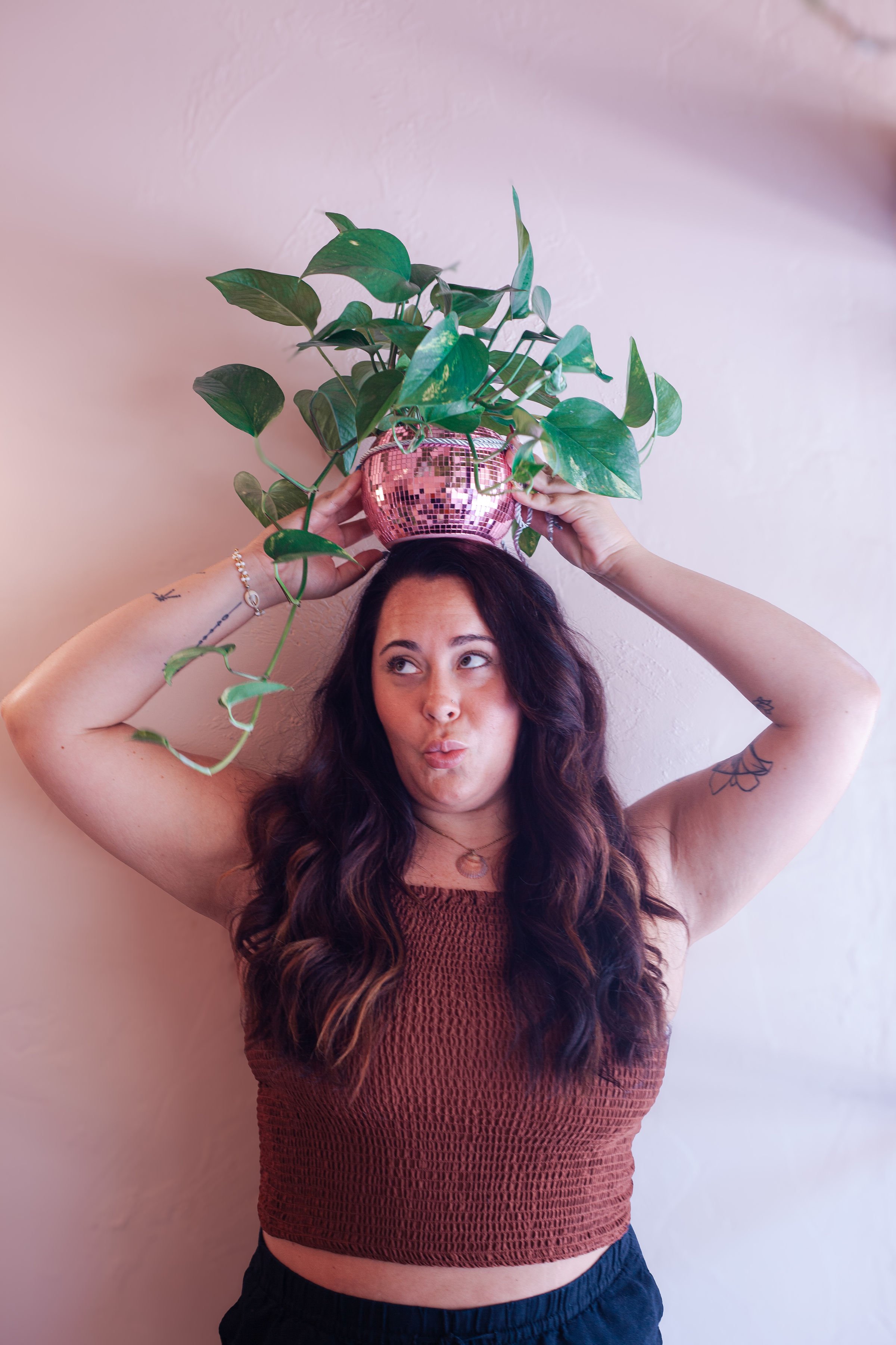 A woman with long wavy dark hair and tattoos on her arms is holding a pink, mirrored disco ball-style vase above her head, with green leafy houseplants cascading out of it. She is wearing a brown sleeveless top and dark shorts, standing against a plain beige wall.