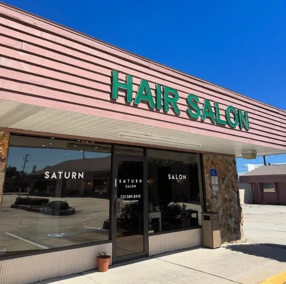 Exterior of a hair salon with pink siding and green signage, with large glass windows and a potted plant outside.