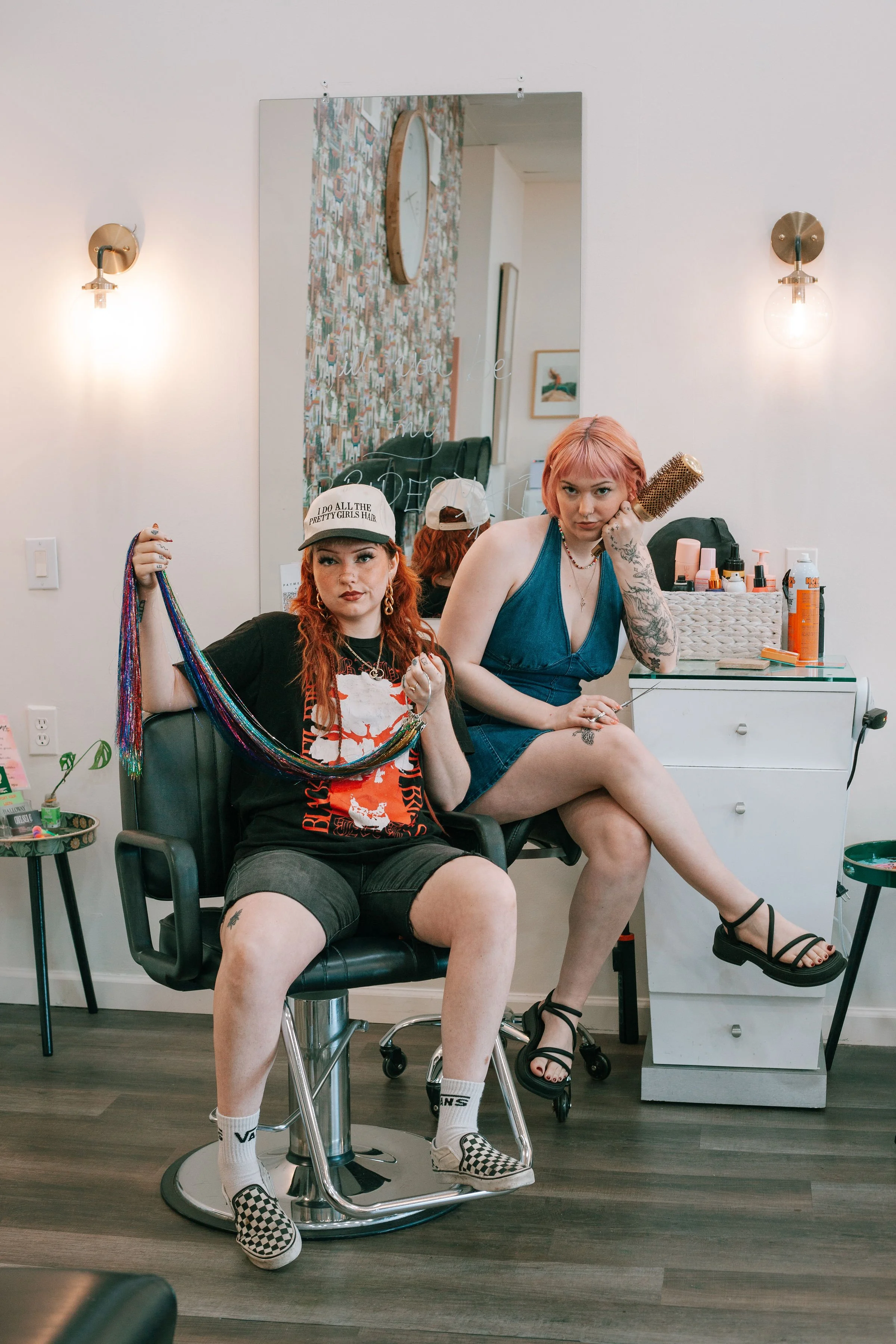 Two women with dyed hair sitting in a salon. One wearing a baseball cap and holding colorful yarn, the other using a round hairbrush. The salon has a mirror, wall-mounted lights, and various hair products on a white drawer unit.