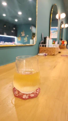 A glass of yellow beverage on a coaster on a wooden counter in a cozy cafe with teal walls, mirrors, and decorative lighting fixtures.