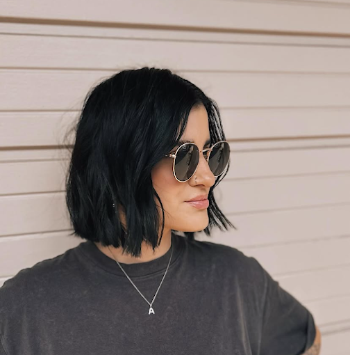 Woman with black, shoulder-length hair wearing sunglasses and a necklace with the letter 'A' against a beige horizontal panel background.