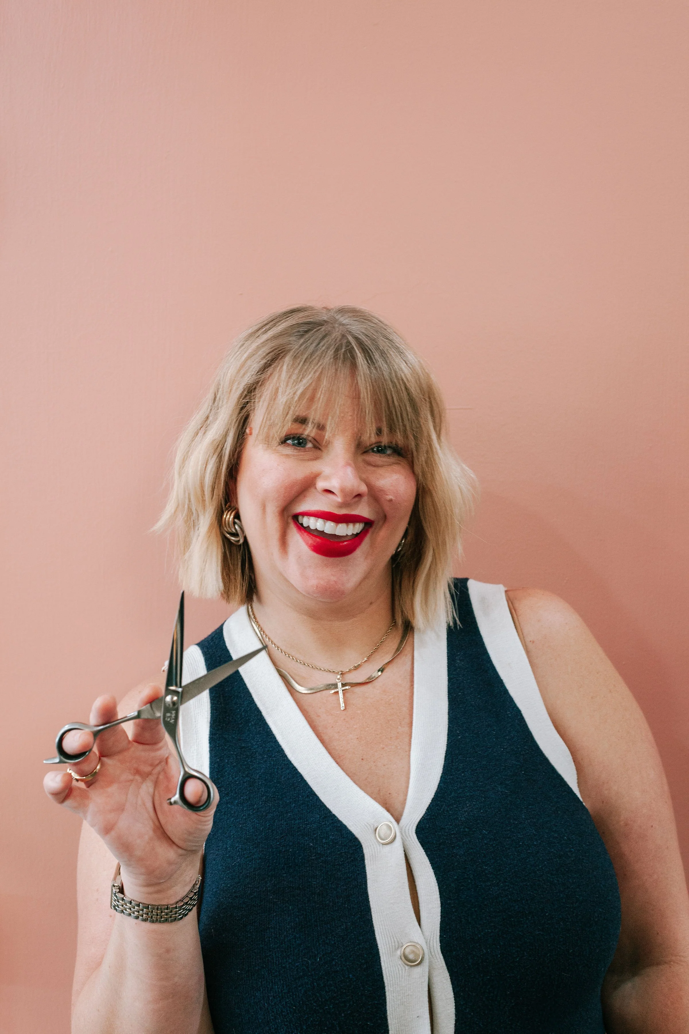 A woman smiling and holding a pair of scissors against a pink background.